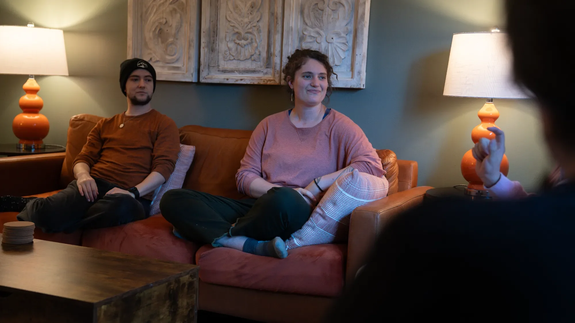 Three people sitting on a brown couch in a living room, two women and one man, engaged in conversation. There are two lamps with orange bases on either side of the couch, colorful wall art behind them, and a wooden coffee table in front.