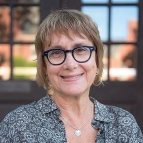 A woman with short, light brown hair wearing glasses and a patterned shirt, smiling outdoors with a building in the background.