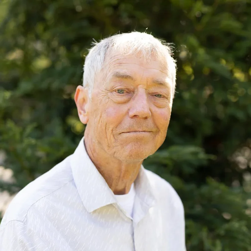 Portrait of an elderly man with white hair, light skin, and a slight smile, wearing a white shirt, standing outdoors in front of green foliage.