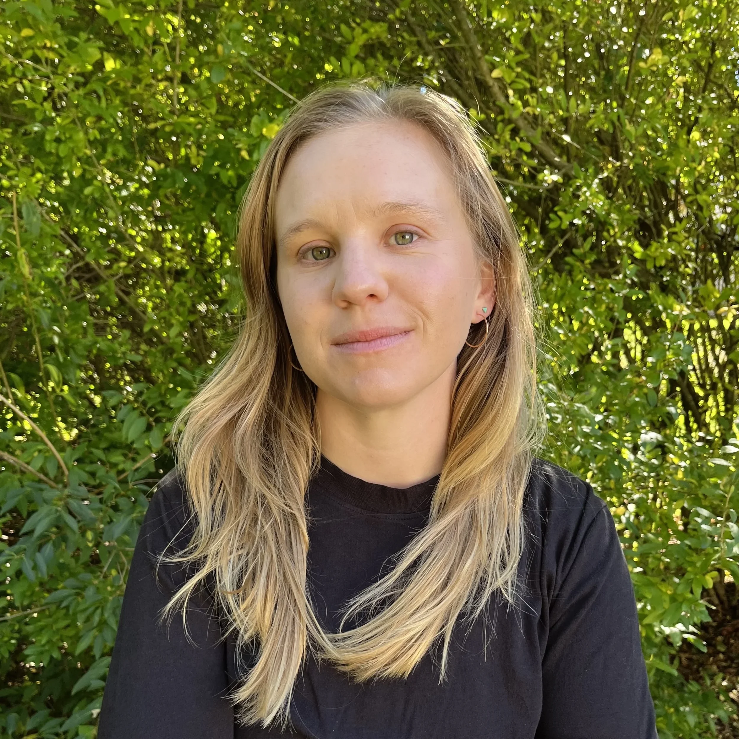 A young woman with light skin, blonde hair, and green eyes stands outdoors in front of green foliage. She is wearing a black shirt and small hoop earrings, with a gentle expression on her face.