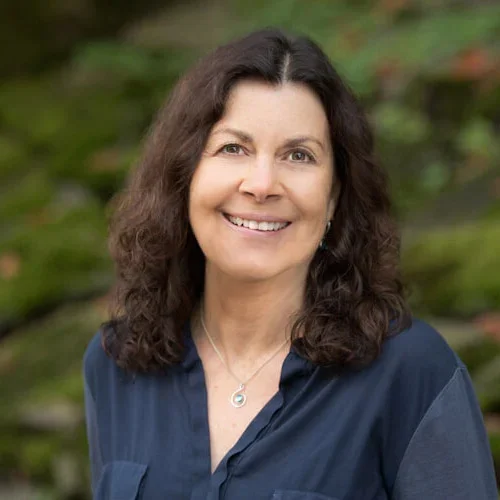 Portrait of a woman with shoulder-length dark hair wearing a dark blue blouse and a silver necklace, smiling outdoors with blurred greenery in the background.