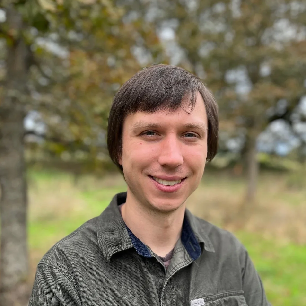 A young man smiling outdoors with trees in the background.