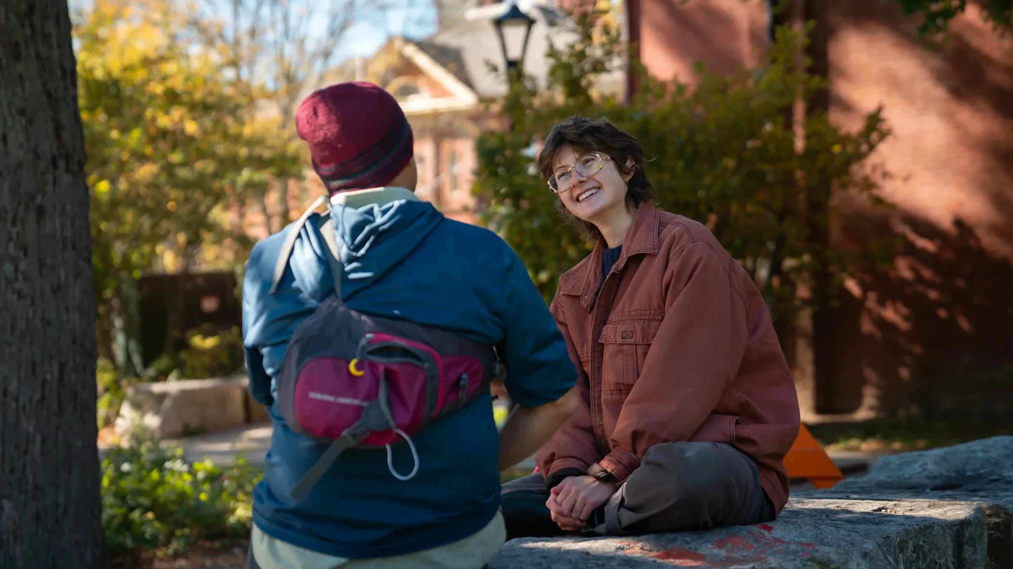 A woman with short dark hair, glasses, and a brown jacket sits cross-legged on a rock, smiling and engaging in conversation with a person facing away, wearing a maroon beanie, blue jacket, and backpack, outdoors with trees and a house in the background.