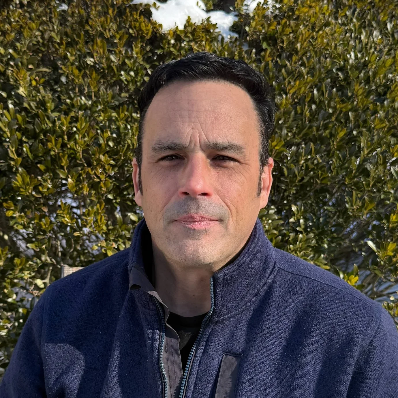 A man with dark hair and a serious expression is outdoors in front of a leafy bush during daytime. He's wearing a blue zip-up jacket.