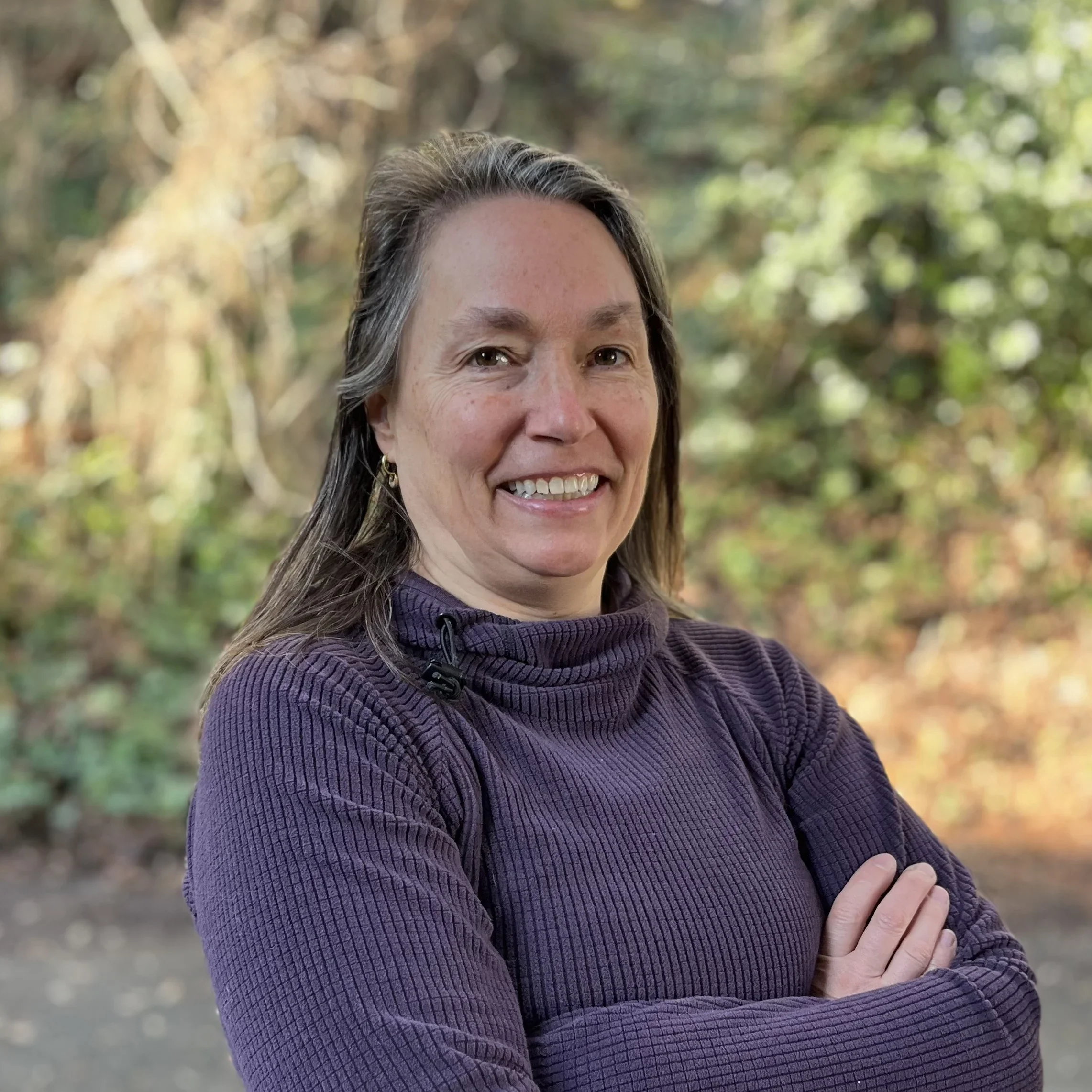 A woman outdoors in a wooded area, smiling with her arms crossed, wearing a purple pullover.