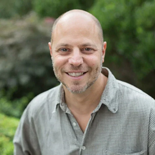 A smiling man with a shaved head and a beard, wearing a light gray checkered shirt, outdoors with green foliage in the background.