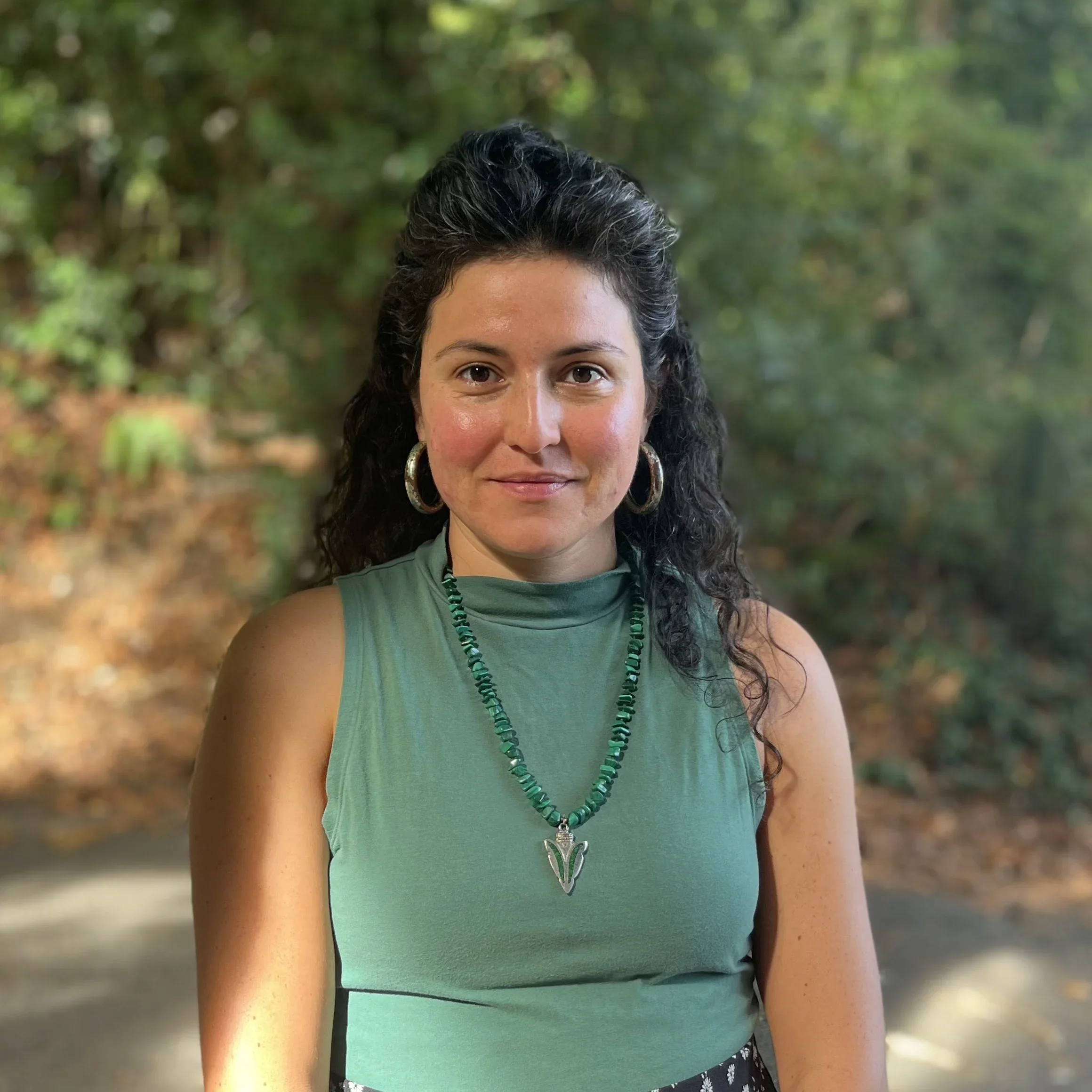 A woman with dark curly hair, wearing large hoop earrings, a green sleeveless top, and a beaded necklace with a pendant, standing outdoors with a blurred green wooded background.