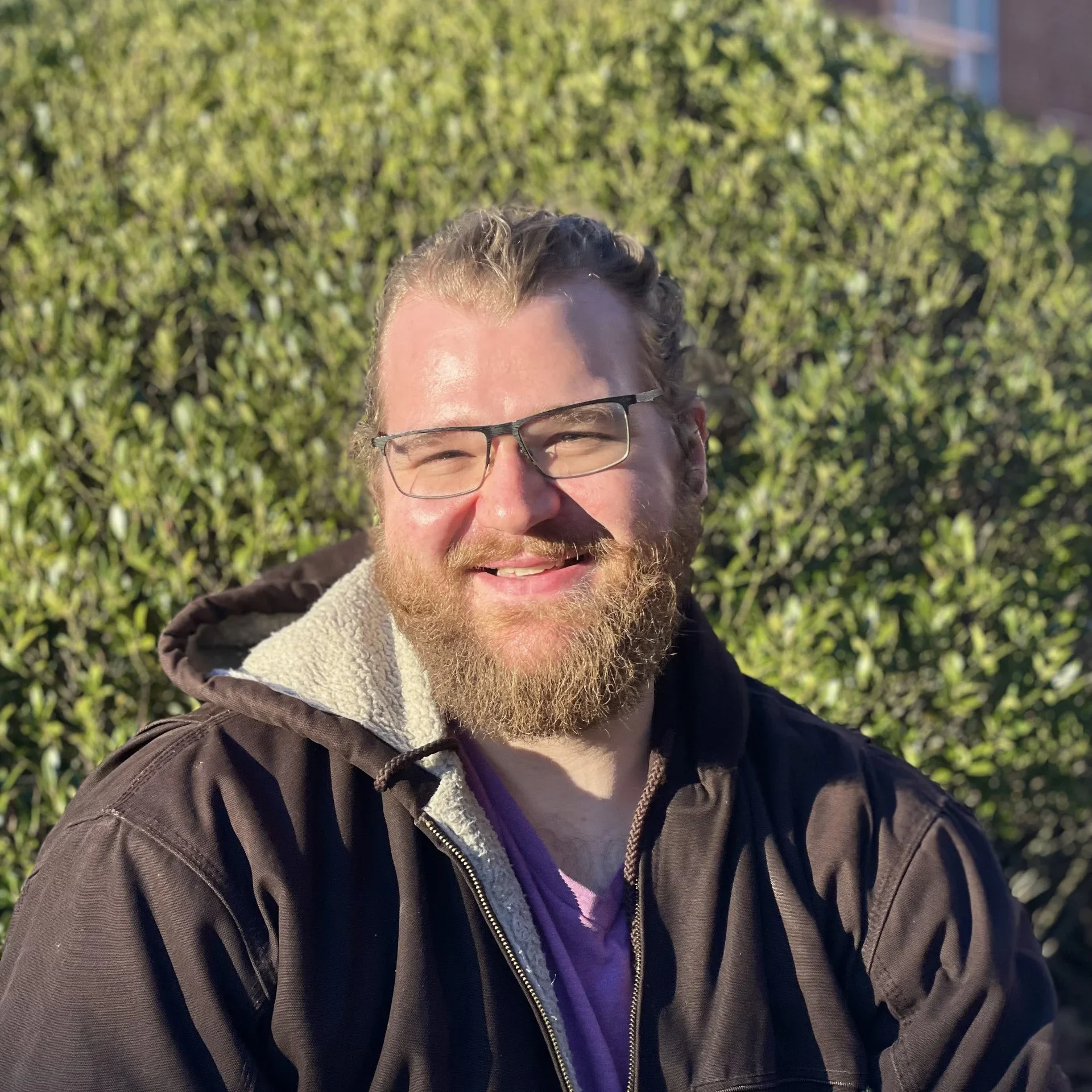 A smiling man with glasses, a beard, and long hair pulled back, wearing a brown jacket with a hood and a purple shirt, outside on a sunny day with greenery in the background.