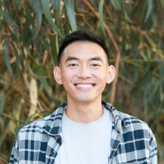 A young man smiling outdoors in front of green foliage, wearing a checkered shirt over a light-colored T-shirt.