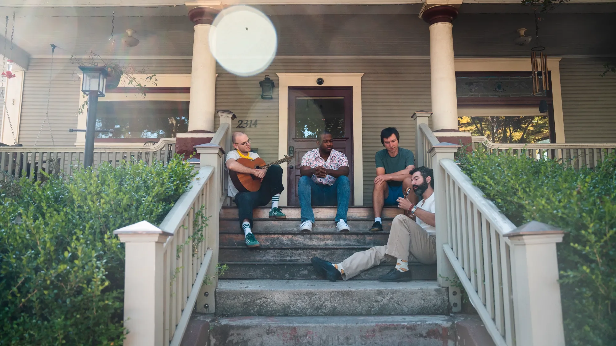 Five men sitting on the front steps of a house, playing guitars, and talking among themselves on a sunny day.