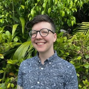 A person with short dark hair, glasses, and a patterned button-up shirt smiling outdoors amid green foliage.
