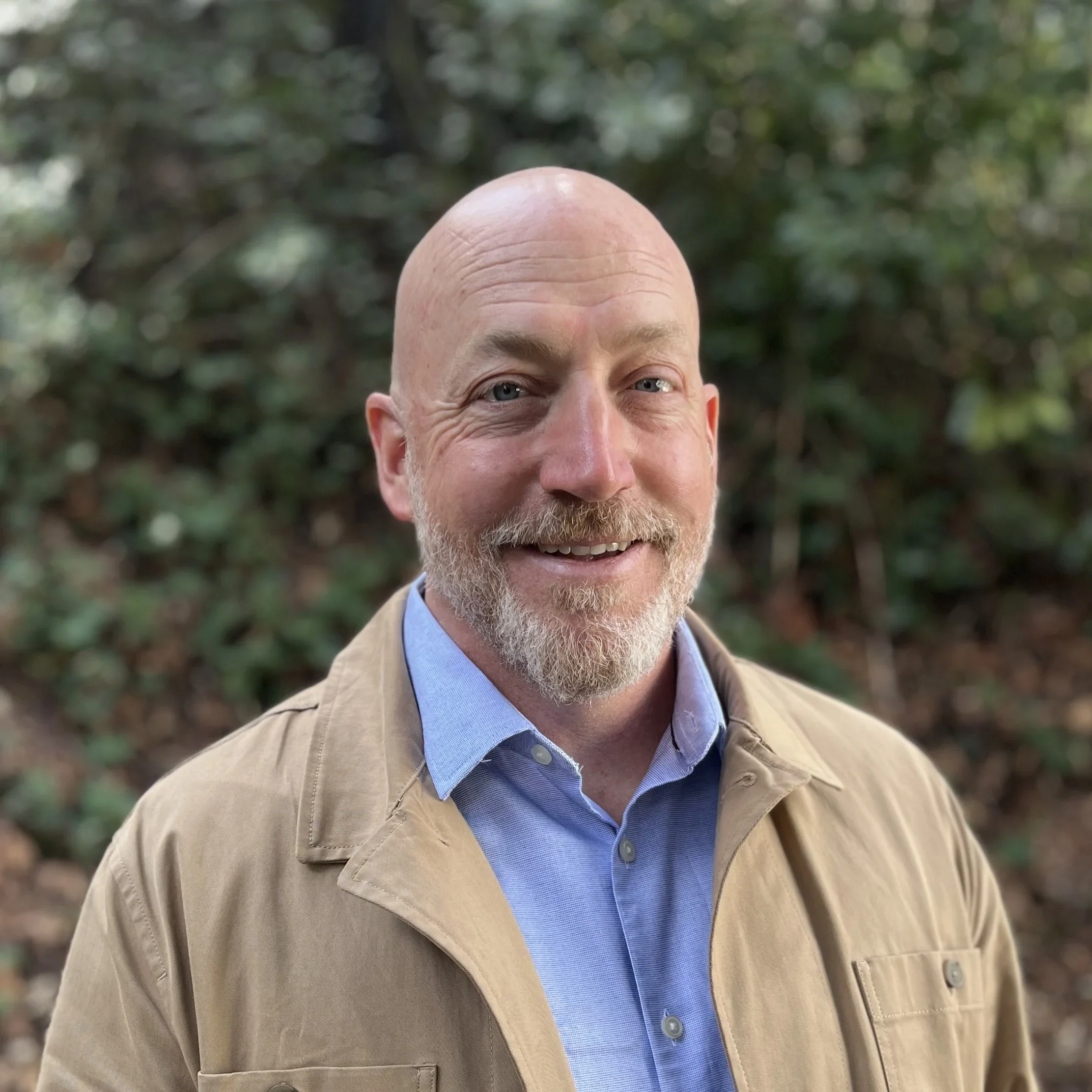A smiling, bald man with a beard wearing a light brown jacket and a light blue shirt, standing outdoors with green foliage in the background.