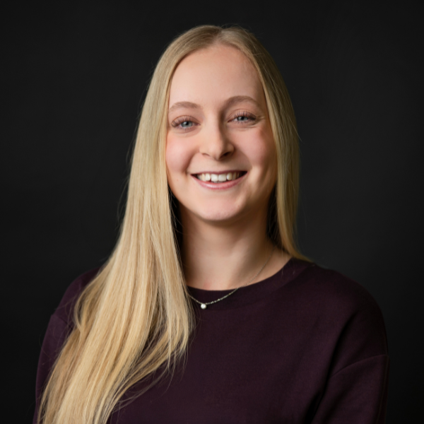 A young woman with long blonde hair, smiling, wearing a black blazer and a necklace, against a dark background with a light blue circular design.