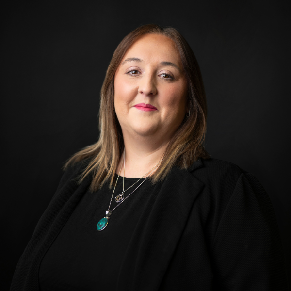 Portrait of a woman with brown curly hair wearing a black blazer and layered necklaces, against a dark gray background with a green circular line design.