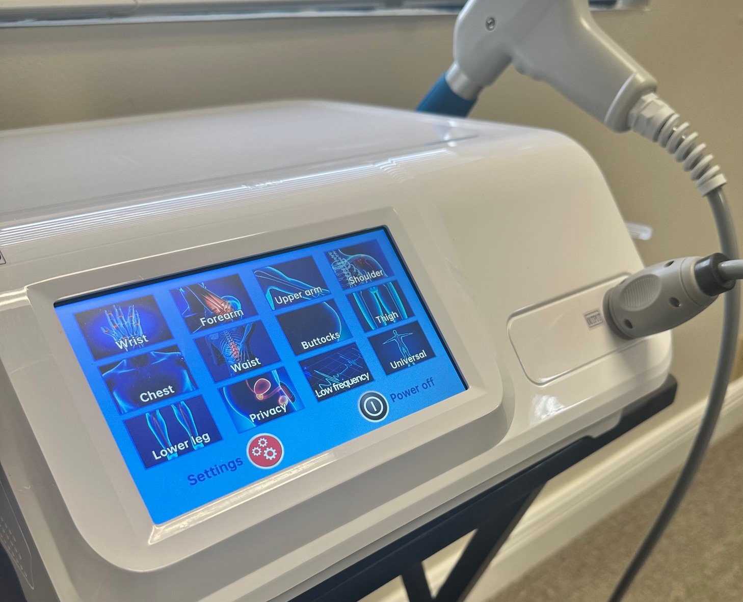 A medical device with a touchscreen display showing options for different body parts, including wrist, forearm, upper arm, shoulder, chest, waist, buttocks, thigh, lower leg, lower leg, and universal. The device has connected attachments.