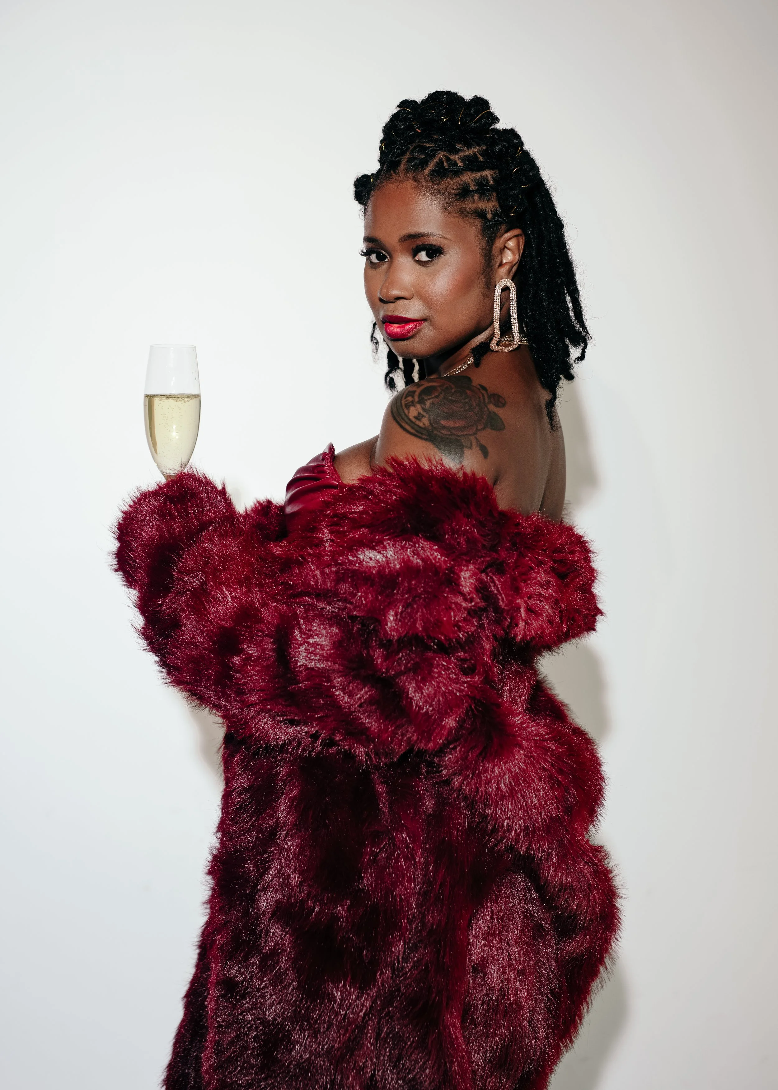 A woman wearing a red fur coat holding a glass of champagne and looking over her shoulder at the camera.