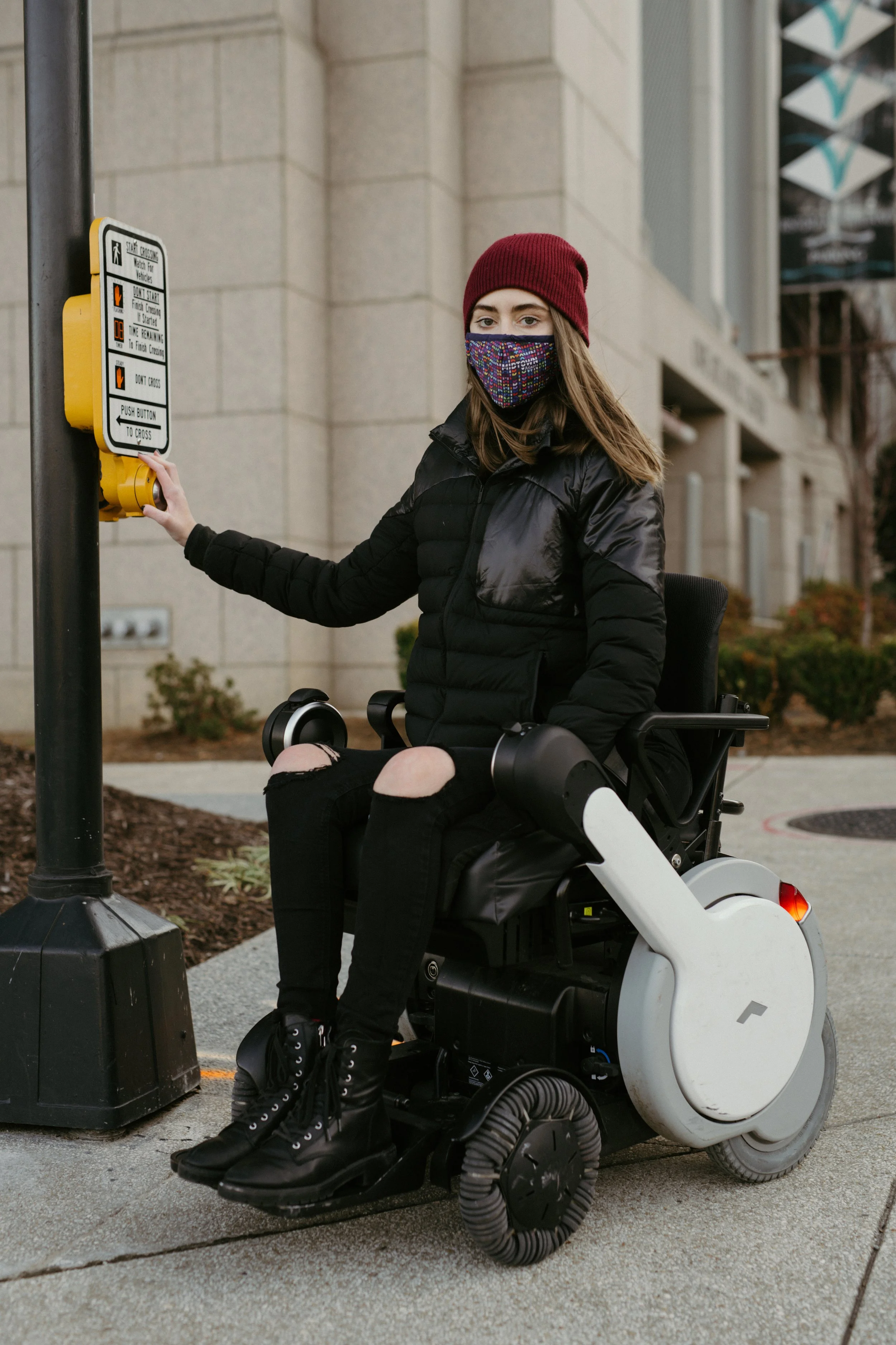 A young woman in a red beanie and colorful face mask operating a crosswalk button while seated in a motorized wheelchair on a city sidewalk.