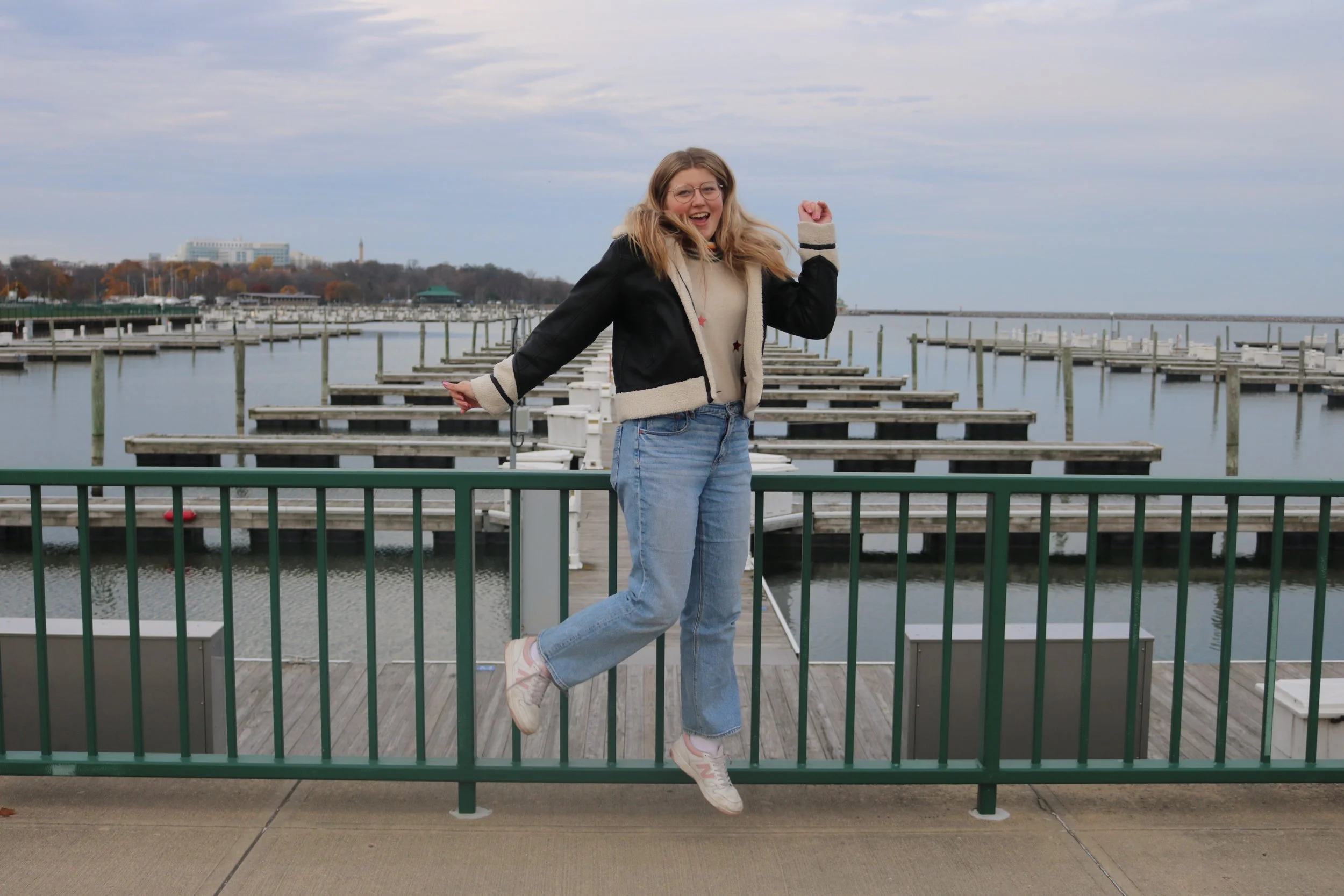A smiling woman jumping with one arm raised on a pier with empty boat docks, overcast sky, and cityscape in the background.