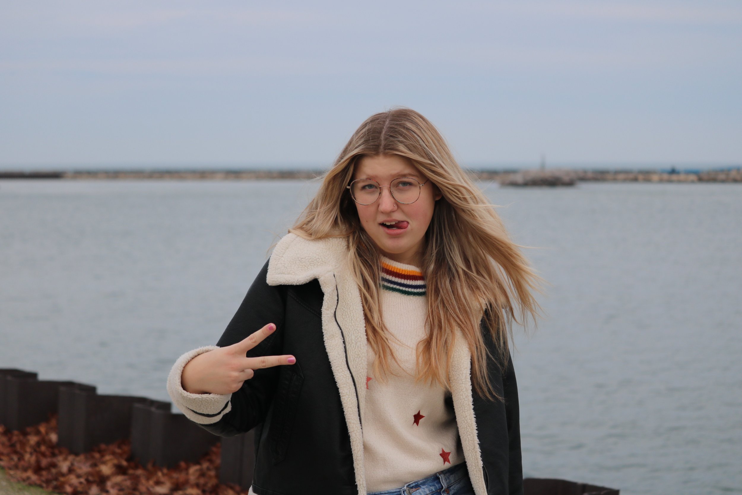A young woman with long blonde hair, glasses, and a beige sweater with red stars making a peace sign and sticking out her tongue by a body of water on a cloudy day.