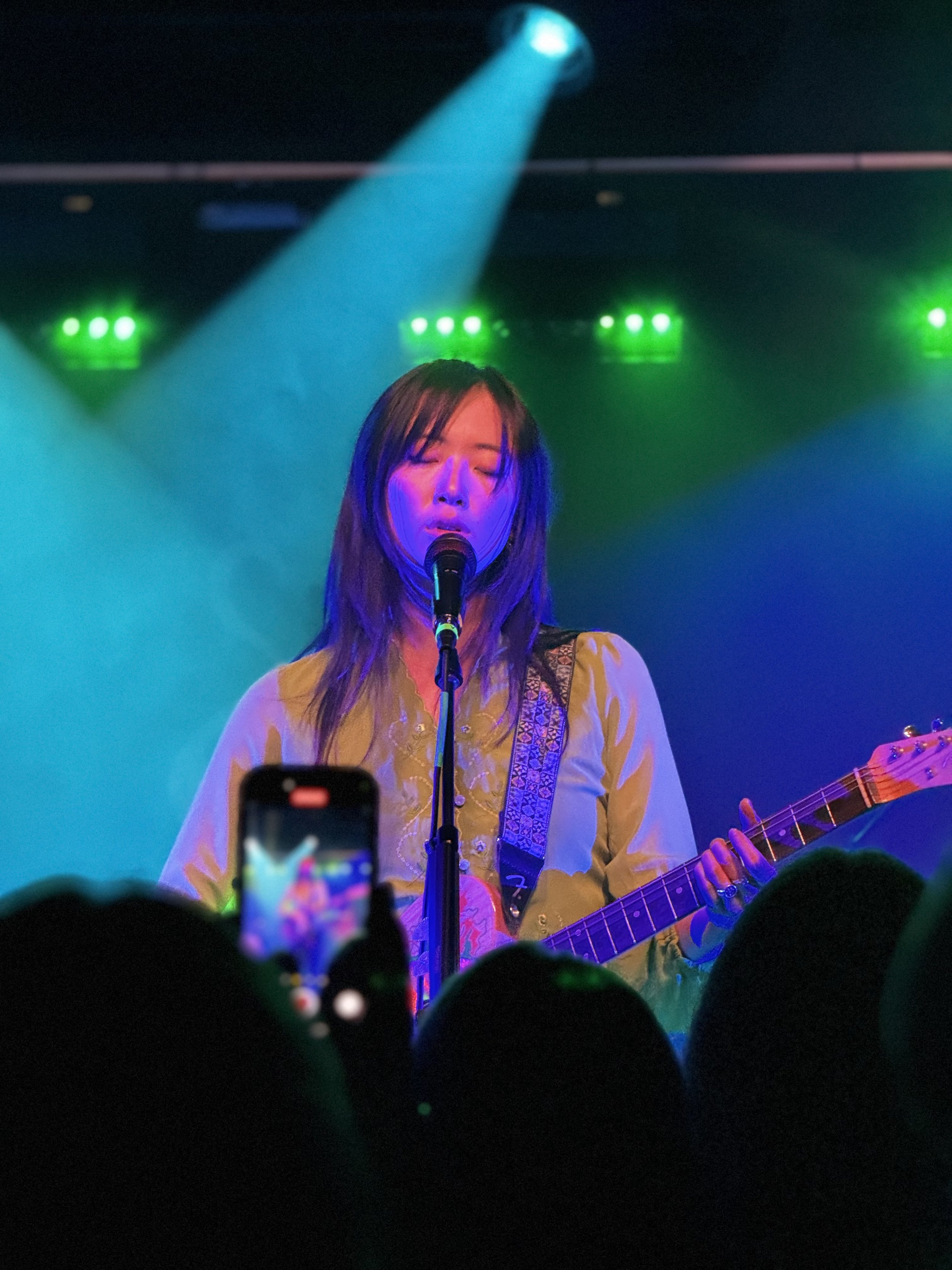 A female singer with long dark hair performs on stage with a guitar, under colorful green and blue stage lights, holding a microphone and singing with closed eyes. An audience member captures the moment on a smartphone.