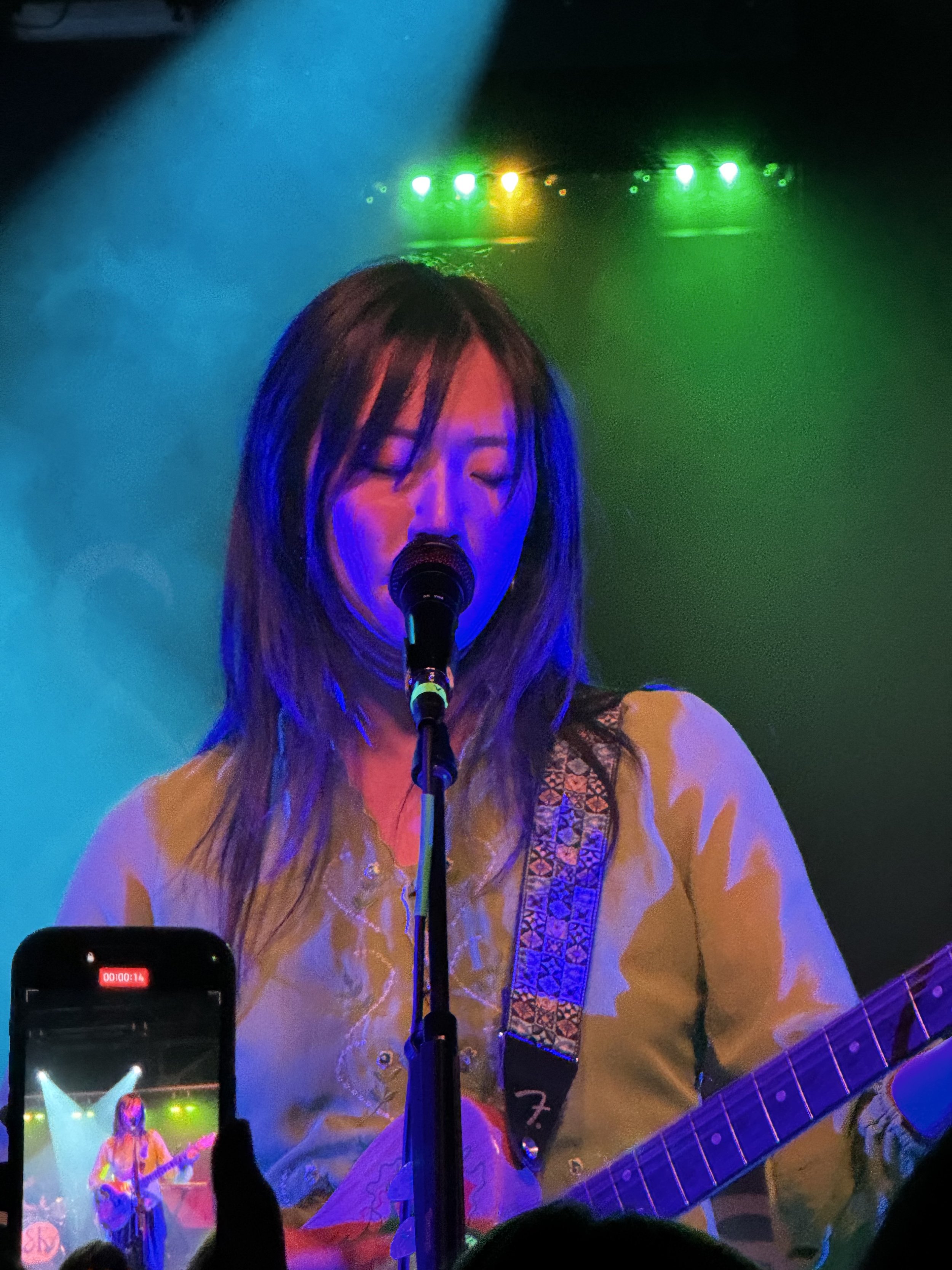 Female musician performing on stage with green and yellow lights, singing into a microphone, playing an electric guitar, wearing a patterned strap, with a smartphone recording the performance.