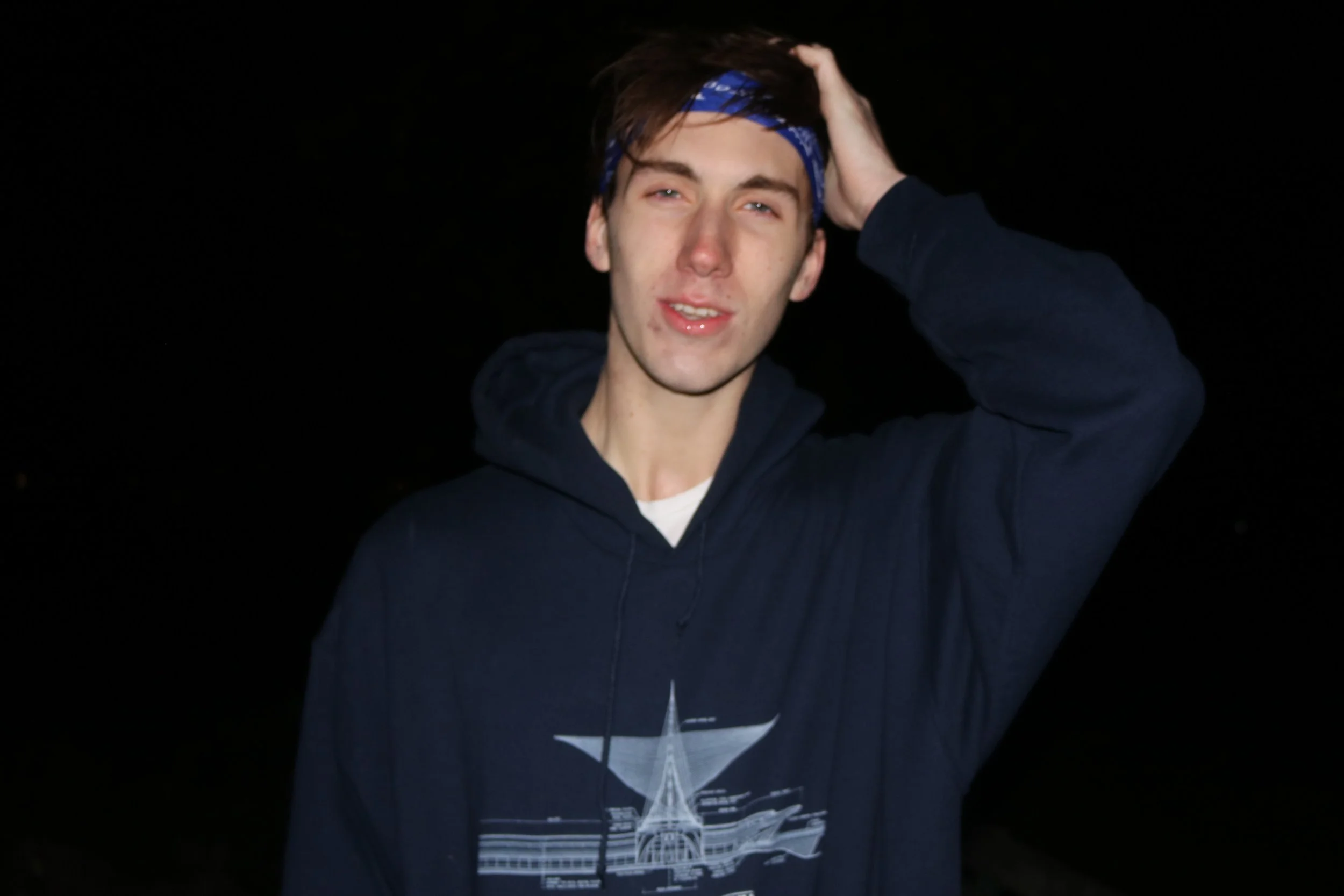 A young man with brown hair, blue eyes, and light skin is wearing a navy blue hoodie with a white graphic, a blue bandana, and is standing against a dark background. He is touching his head with one hand and looking at the camera.