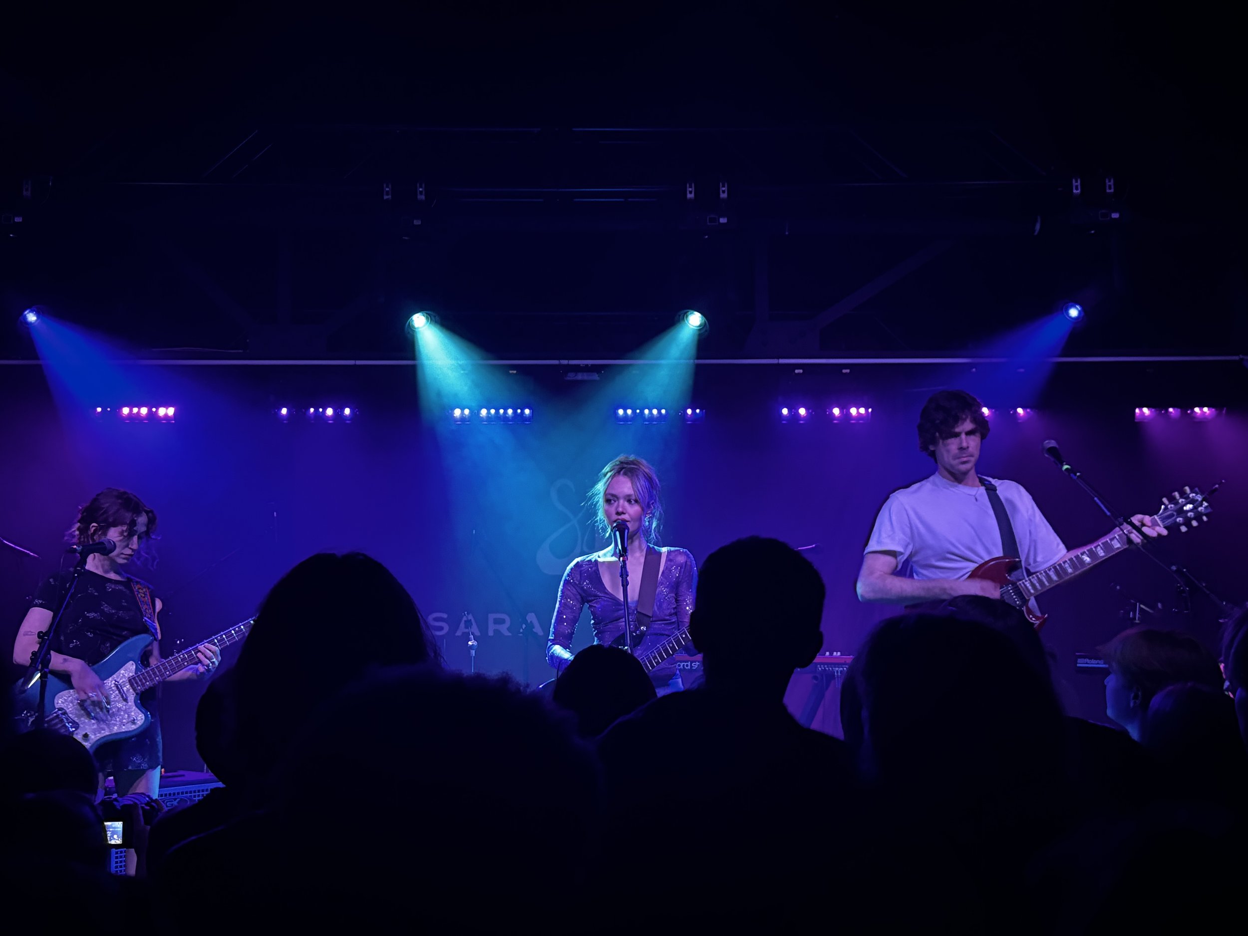 A band performing on stage with three members, illuminated by colorful stage lights, in front of an audience.