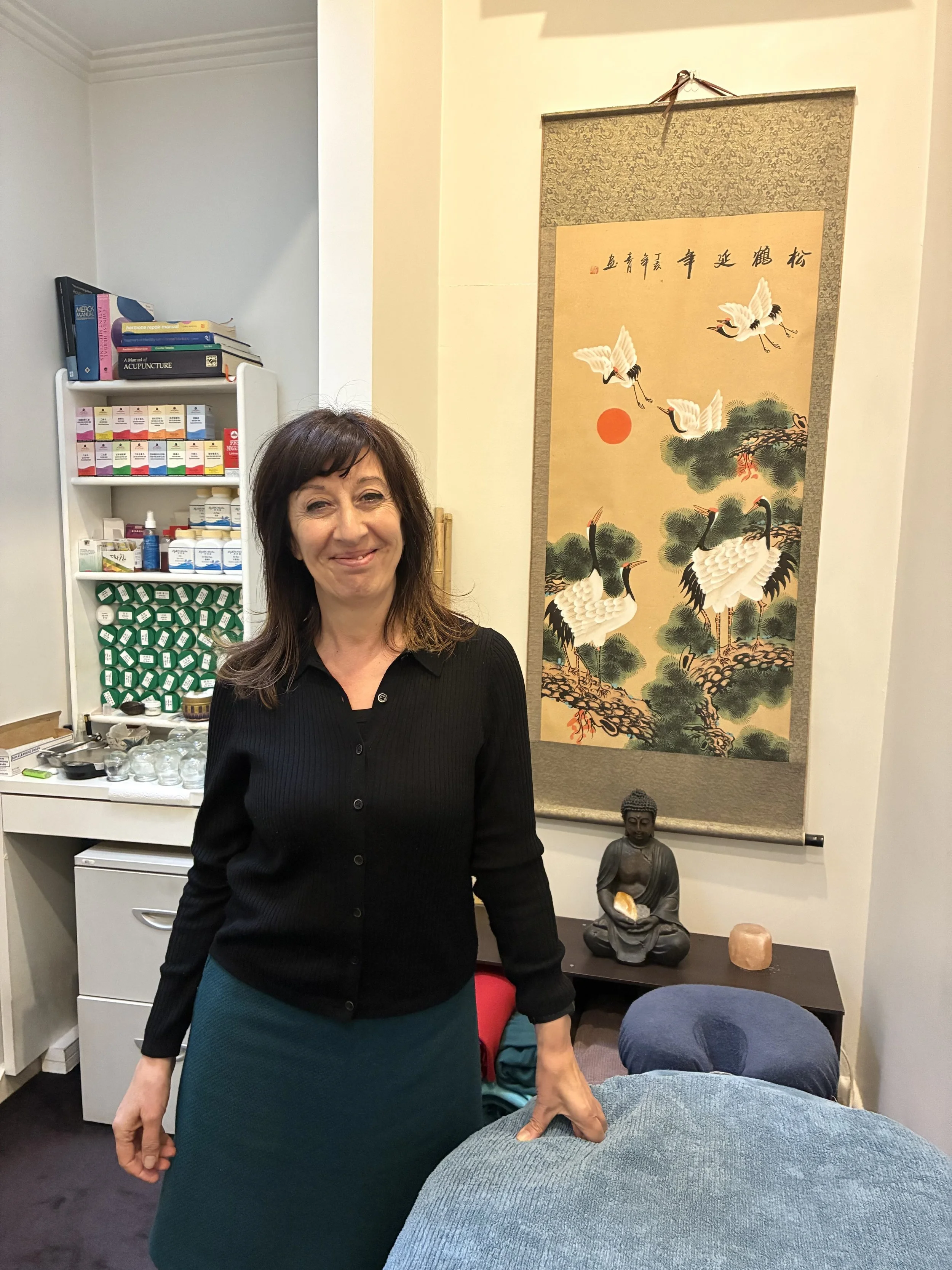 A woman with dark brown hair in a black cardigan and teal skirt standing in an Acupuncture treatment room with Asian-inspired decorations, including a hanging scroll and a Buddha statue, smiling at the camera.
