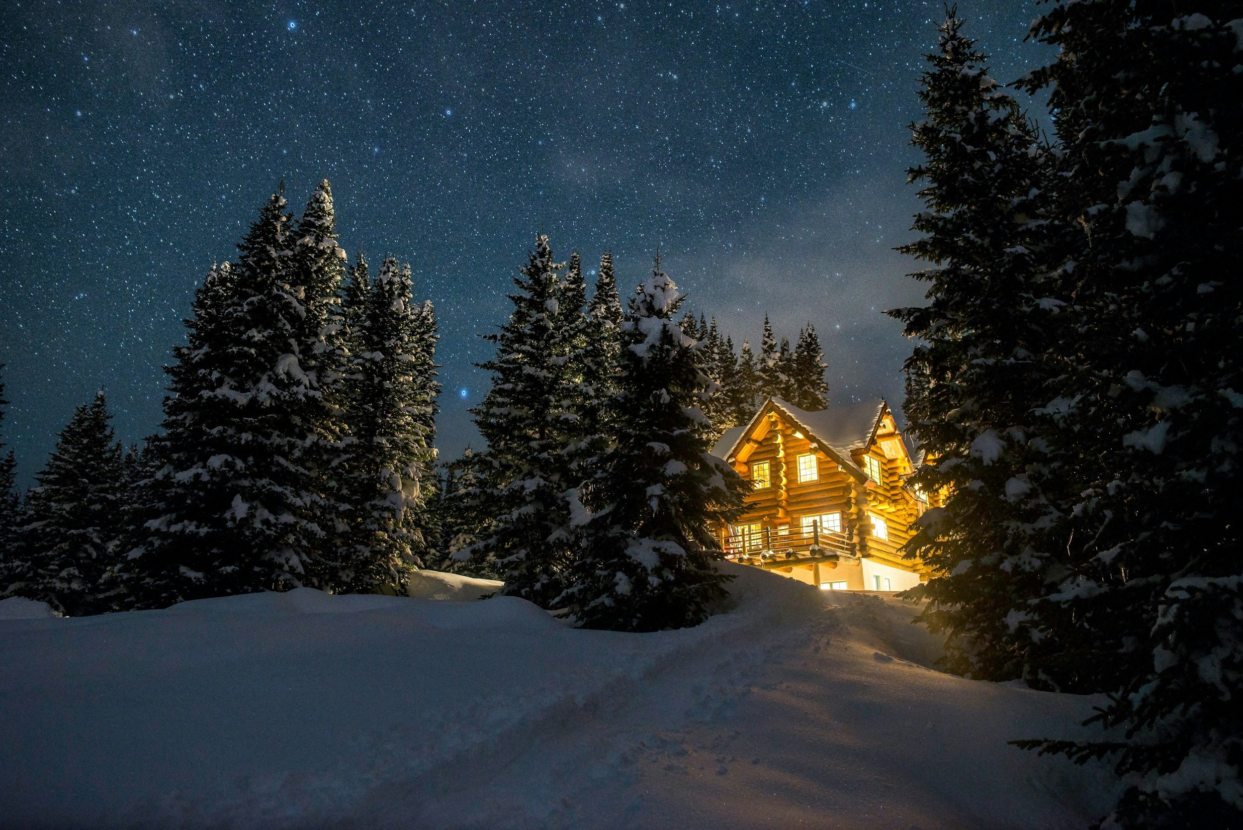 A cozy cabin with warm yellow lights glowing inside, nestled among snow-covered evergreen trees in a winter forest at night, under a starry sky.