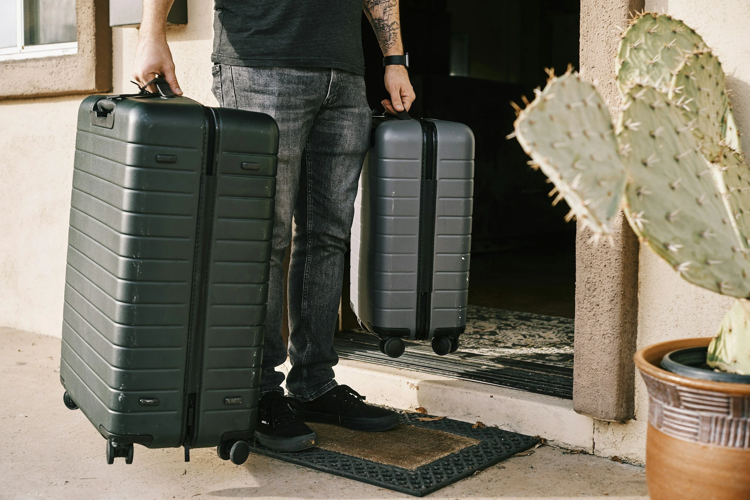 Person standing at a doorway holding two rolling suitcases, one black and one gray, with a cactus plant nearby.