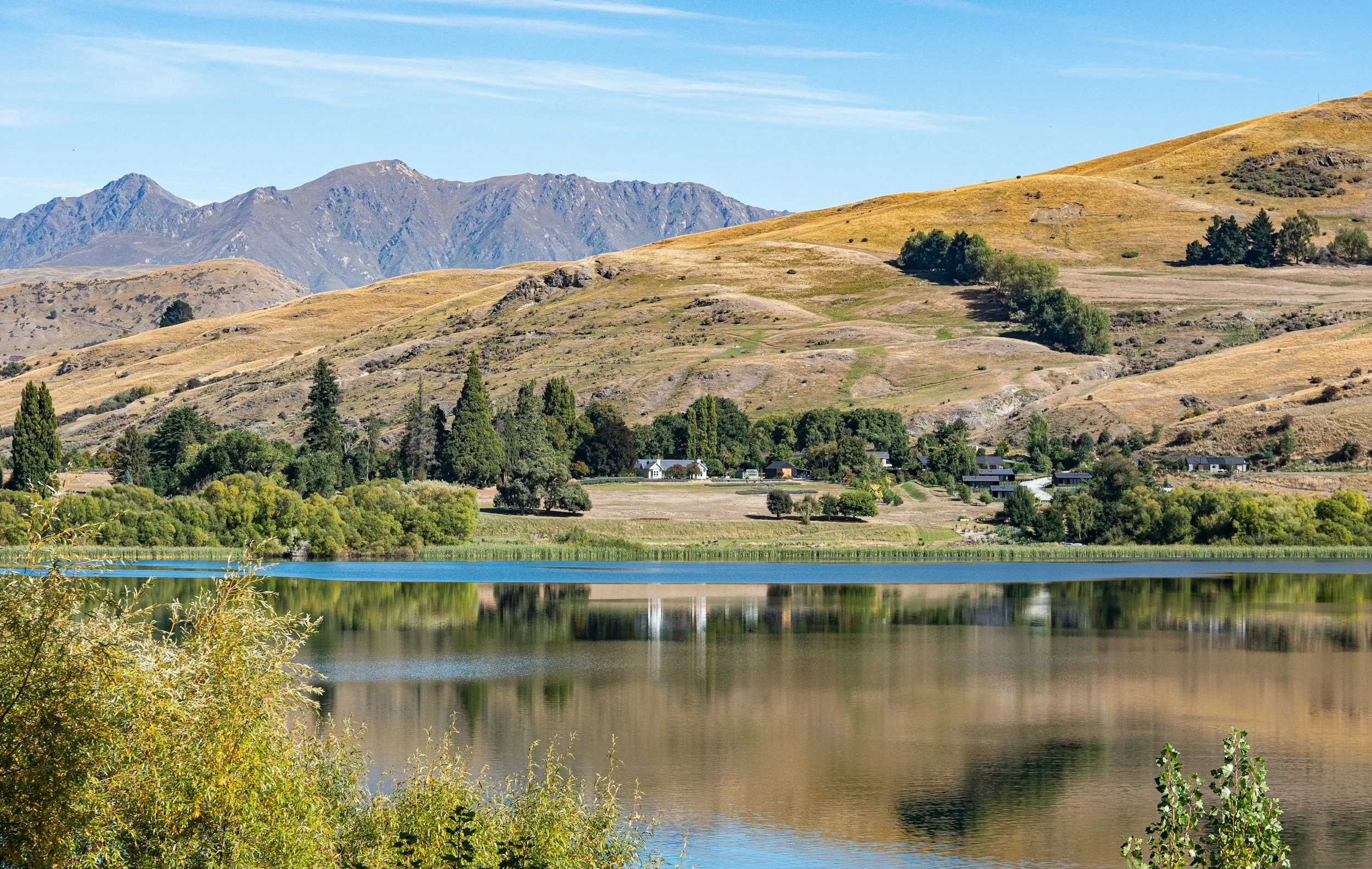 Scenic landscape with a river in the foreground, green trees, a small residential area, rolling hills, and mountains in the background under a clear blue sky.