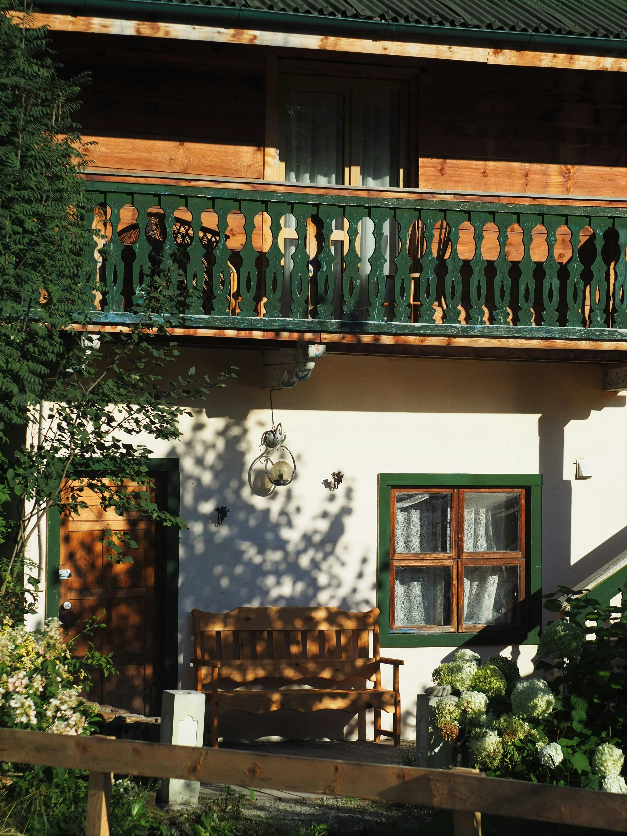 A rustic house with a wooden balcony and a bench outside. The house has white walls, green window frames, and a wooden door. There are plants and flowers in the garden, with a lamp hanging on the wall. Shadows from the trees are cast on the house.