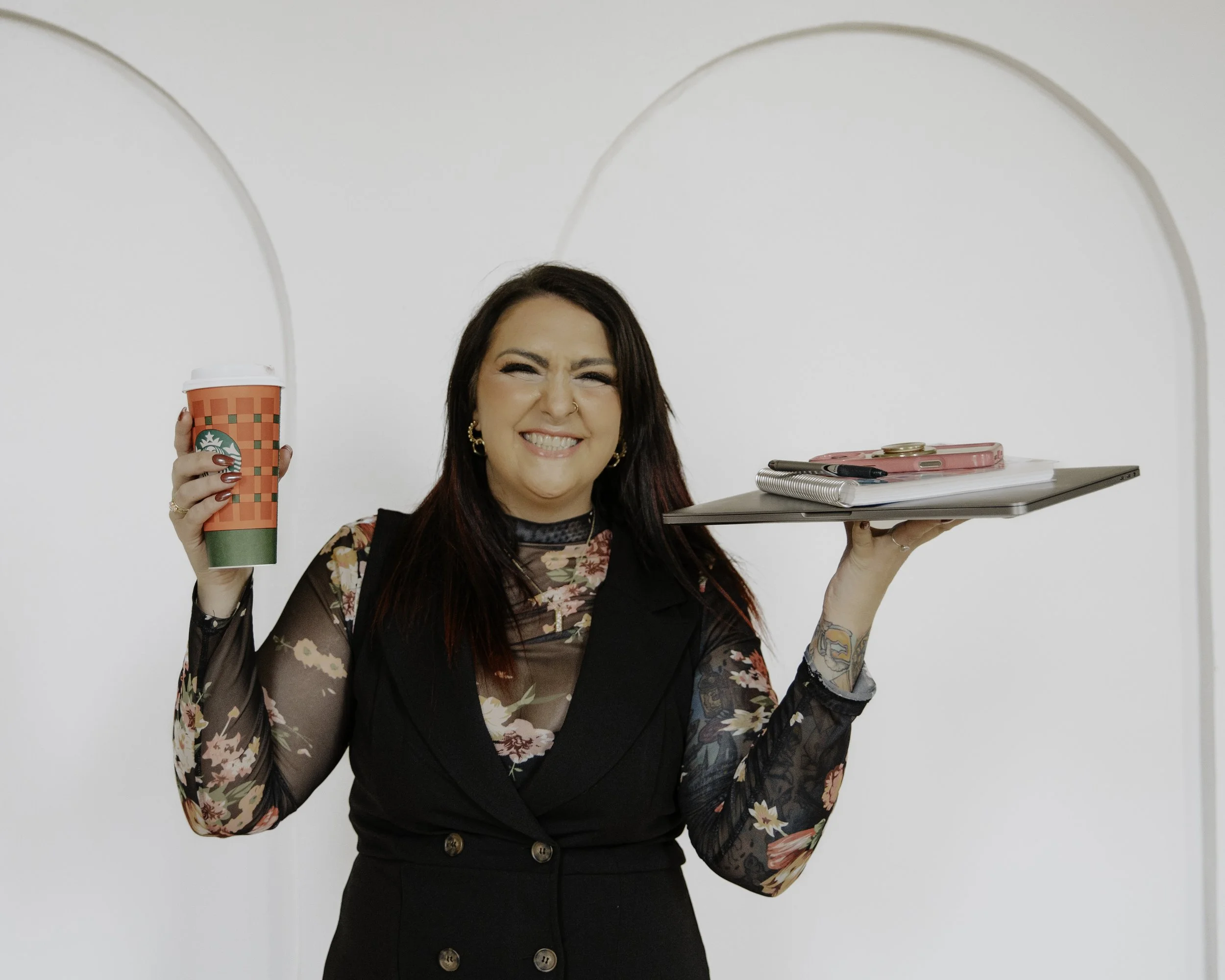 A woman holding a Starbucks holiday-themed cup in one hand and a tray with notebooks, pens, and a coin in the other, smiling brightly against a white wall background.