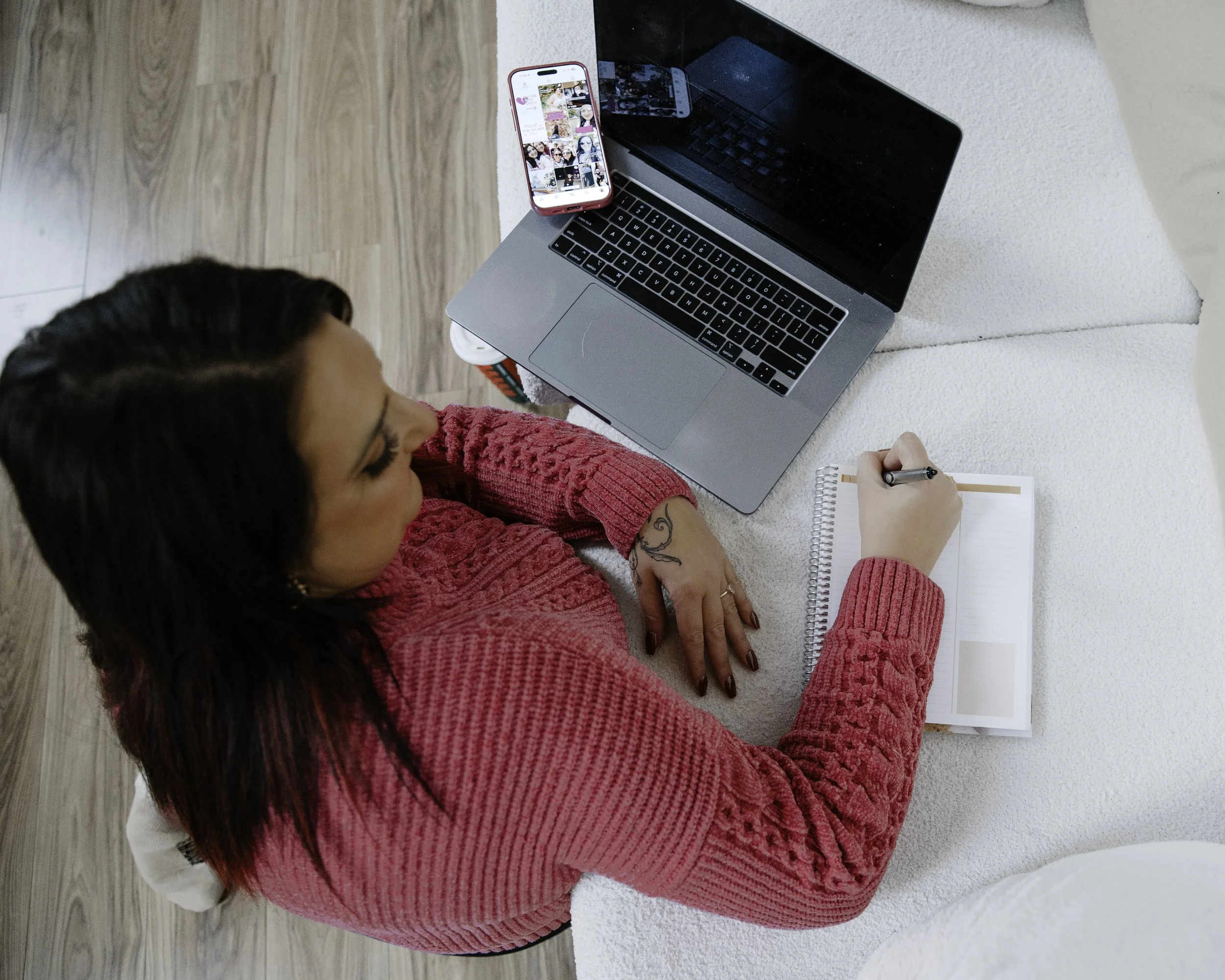 A woman with dark hair wearing a pink sweater, sitting at a white table with a laptop, a smartphone, and a notepad, writing with a pen.