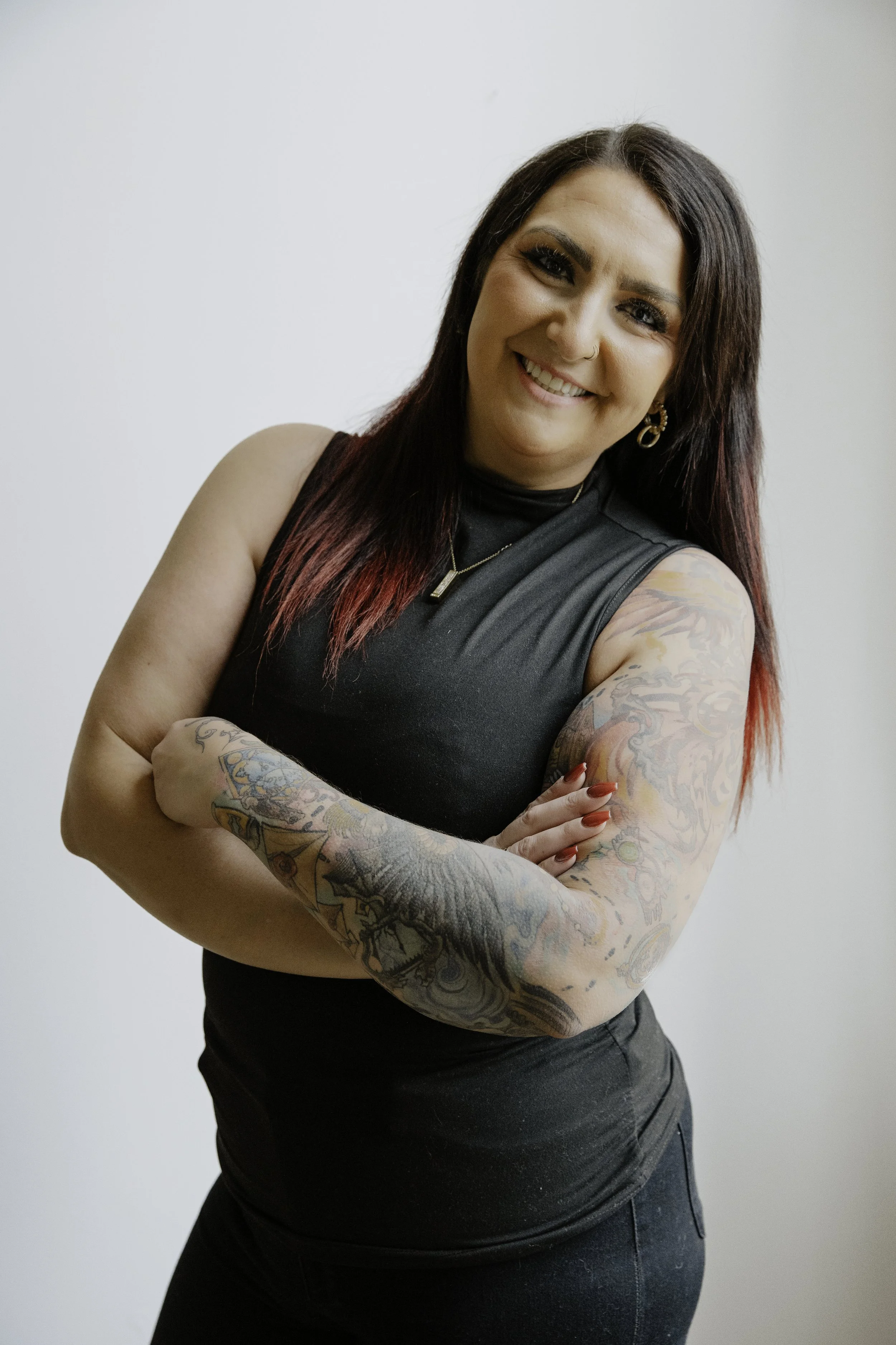 A woman with long dark hair with red highlights, smiling and crossing her arms, wearing a black sleeveless top and showing colorful tattoos on her arms.