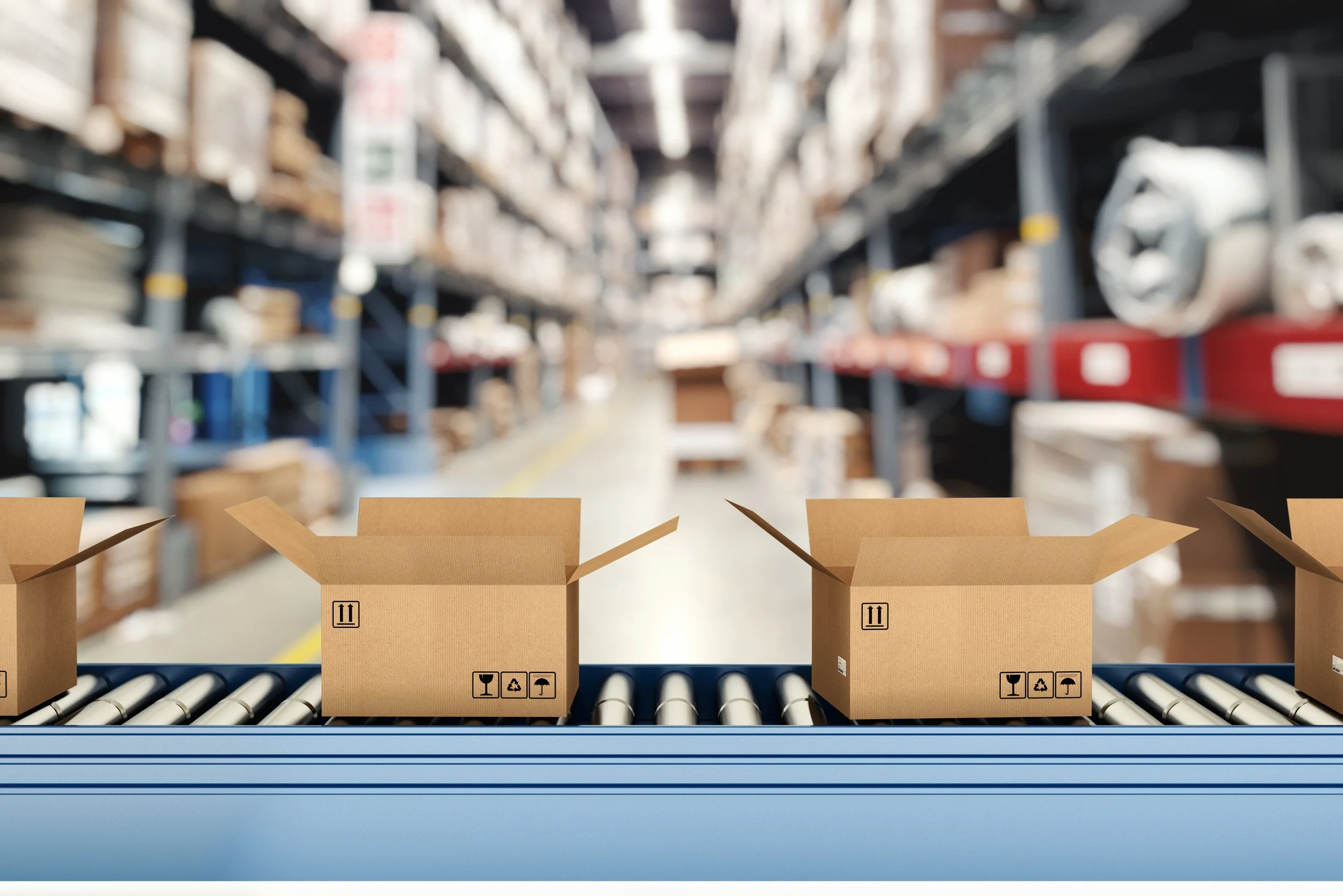 A row of open corrugated cardboard boxes marked with standard handling symbols — fragile, recyclable, and this-side-up — move along a blue roller conveyor belt in the foreground of a large warehouse facility.