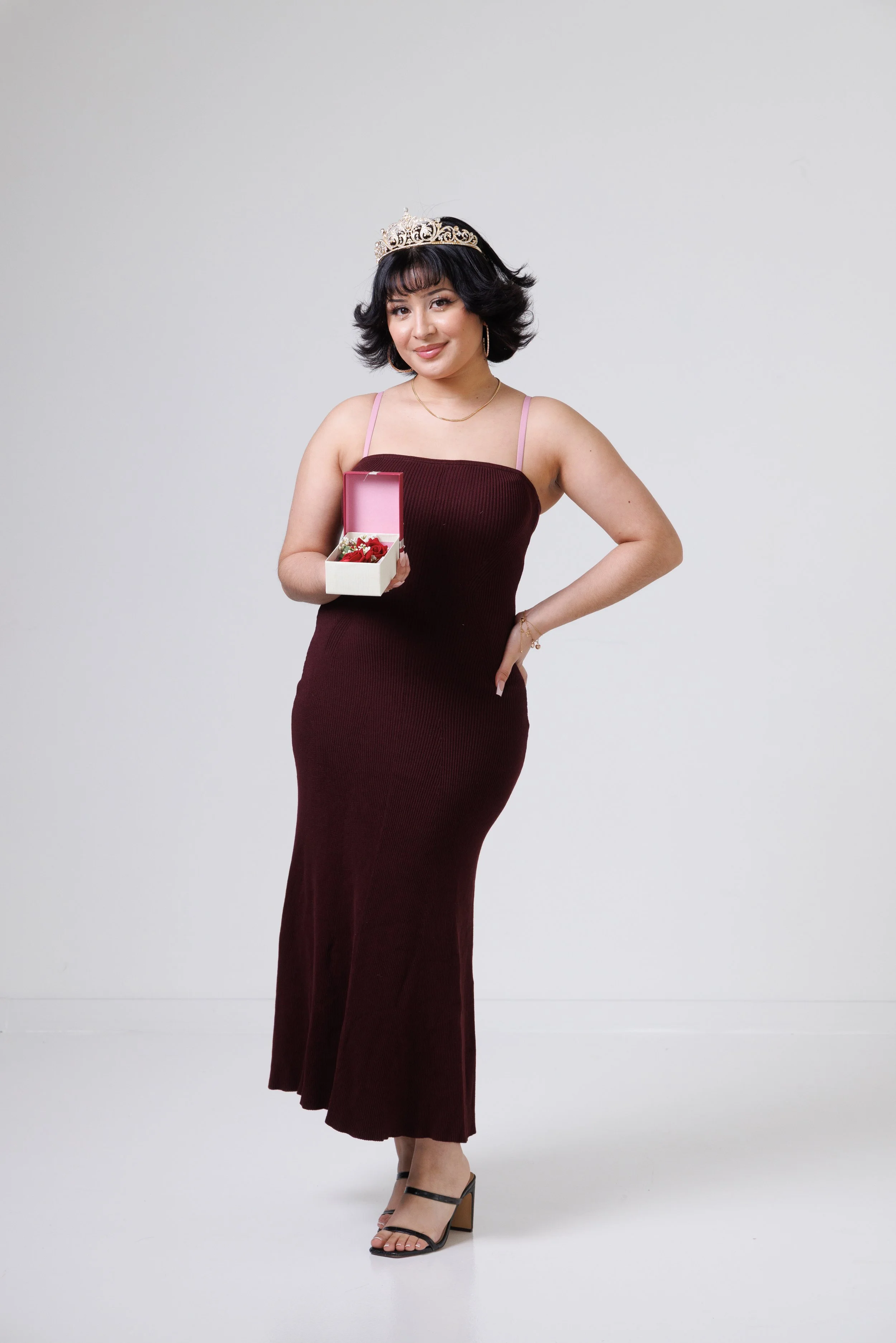 A woman with short black hair wearing a tiara, a burgundy dress, and high-heeled sandals, holding a small box with a red bow against a plain gray background.