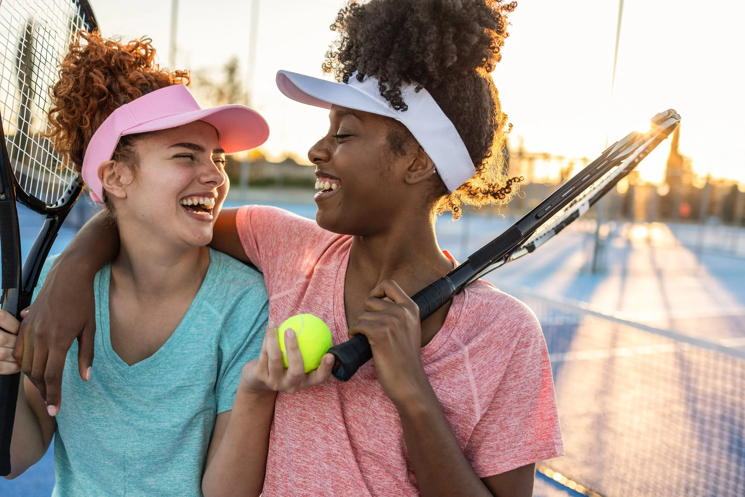 Two young women smiling and laughing on a tennis court during sunset, one holding a tennis ball and the other holding a tennis racket.