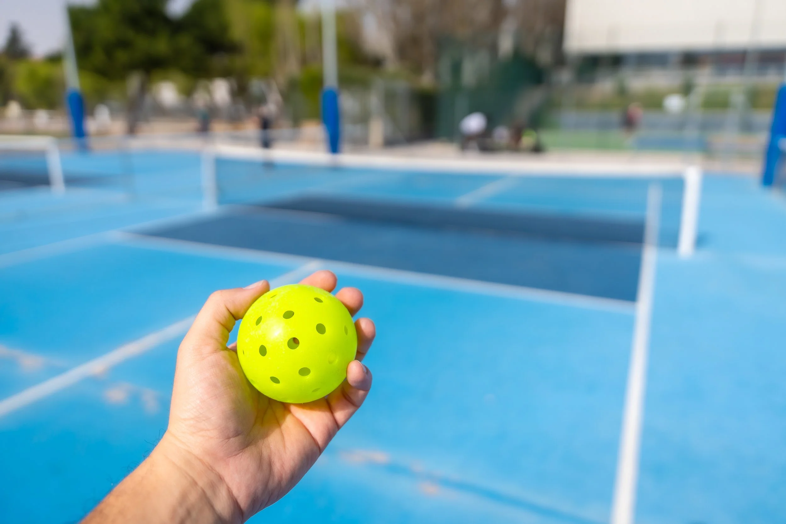 A person holding a yellow perforated tennis ball in front of an outdoor tennis court with blue surface and a net.