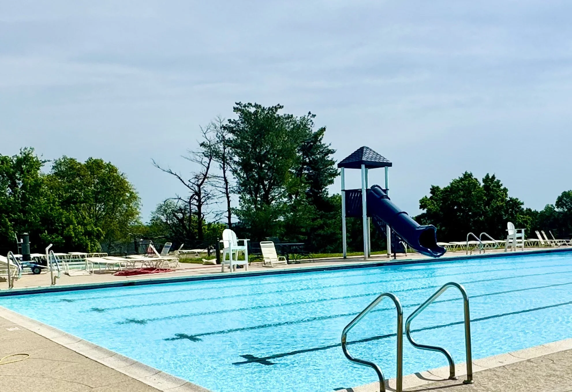 Empty outdoor swimming pool with a slide and lounge chairs, surrounded by trees under a cloudy sky.