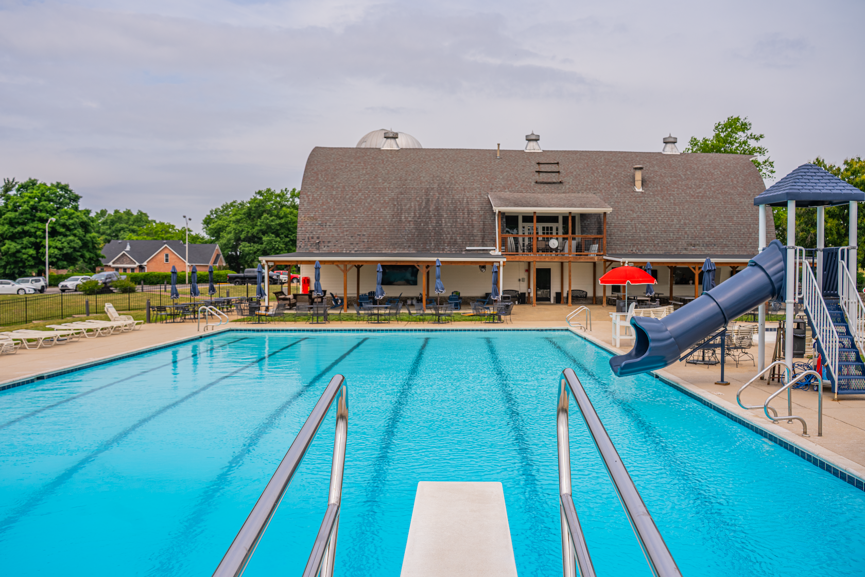 Public swimming pool with blue water, metal handrails, and a slide, surrounded by lounge chairs, tables with umbrellas, and a large building in the background with a deck and classroom.