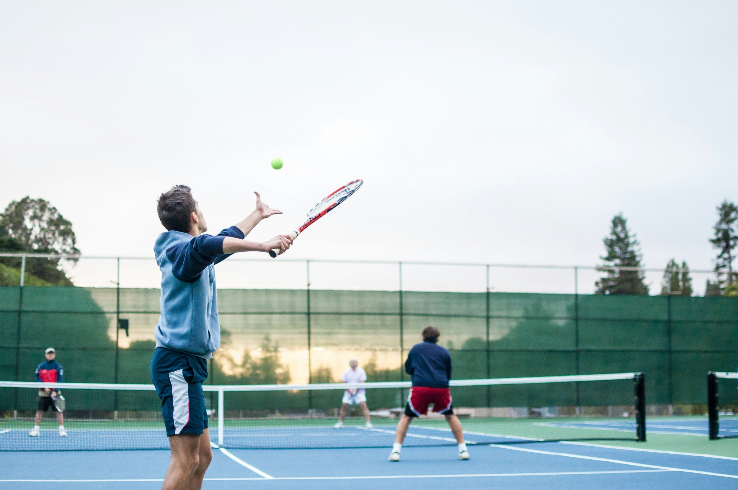 Young boy playing tennis on an outdoor court with other players in the background, overcast sky, surrounded by trees and fencing.
