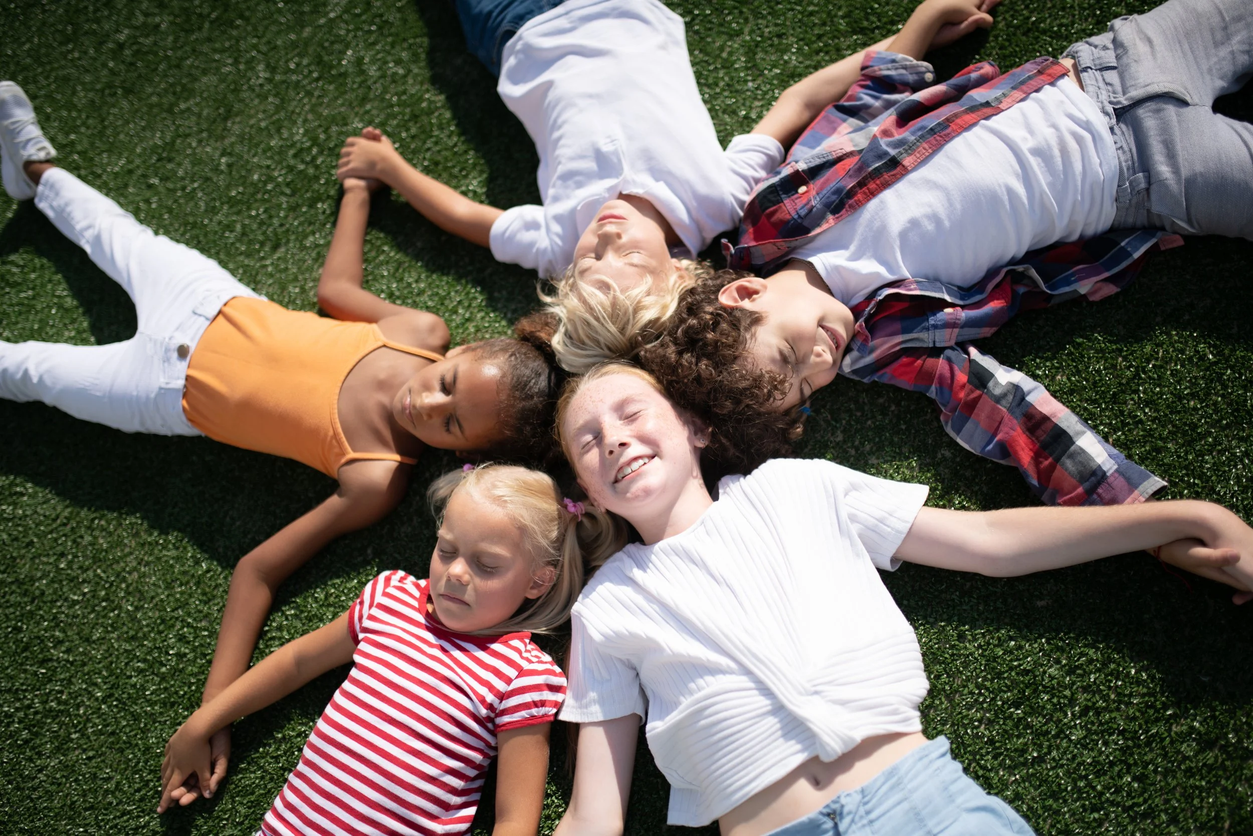 Five children lying on grass in a circle, holding hands and smiling.
