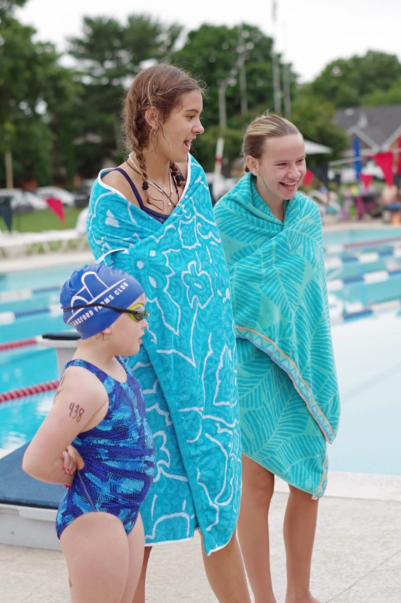 Three female swimmers, one young girl and two possibly teens or young adults, standing beside a swimming pool after a race, wrapped in blue towels, with the pool and other swimmers visible in the background.