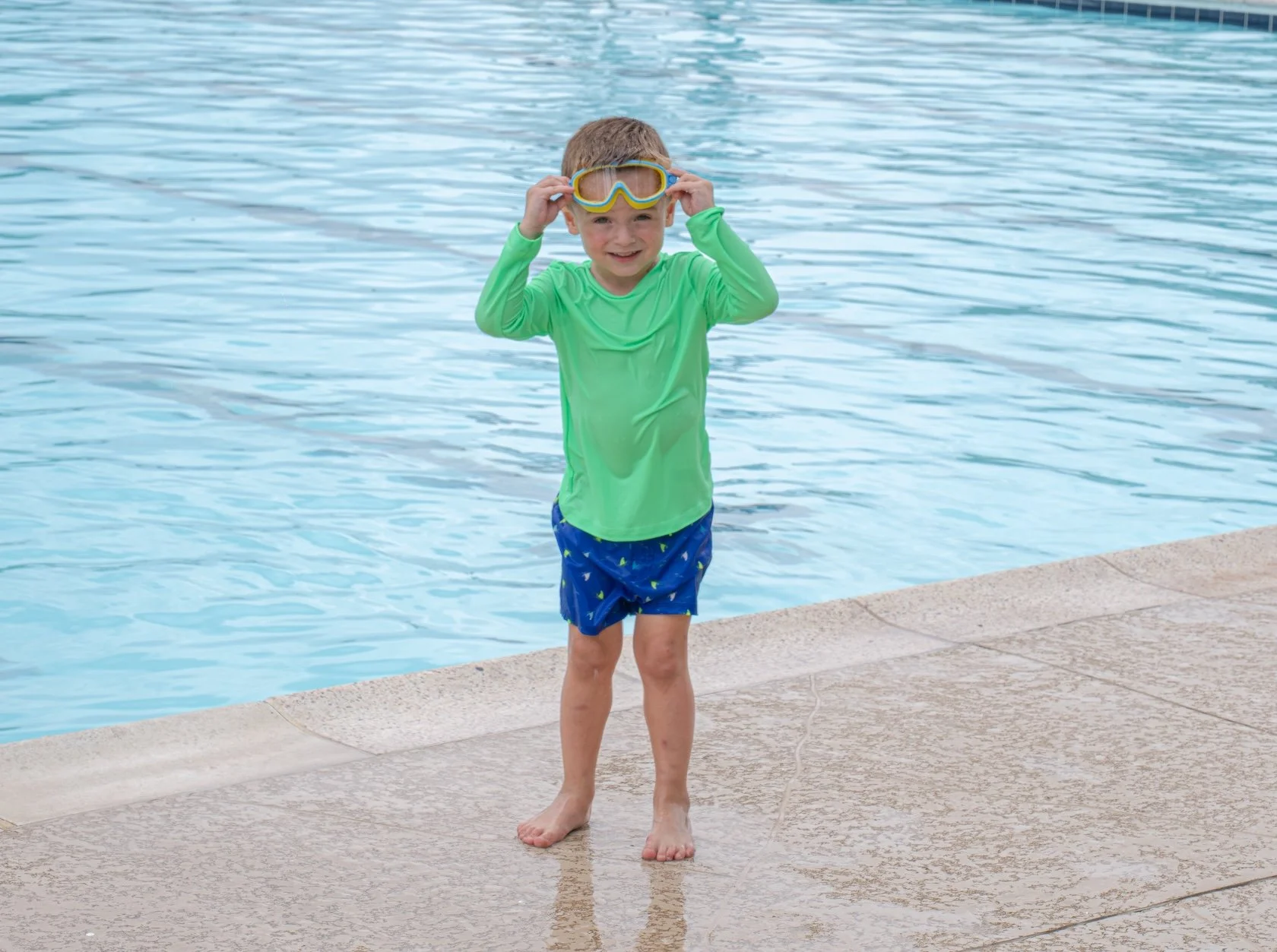 Young boy standing at the edge of a swimming pool, wearing a green long-sleeve swim shirt, blue swim trunks with a pattern, and yellow swimming goggles, smiling at the camera.