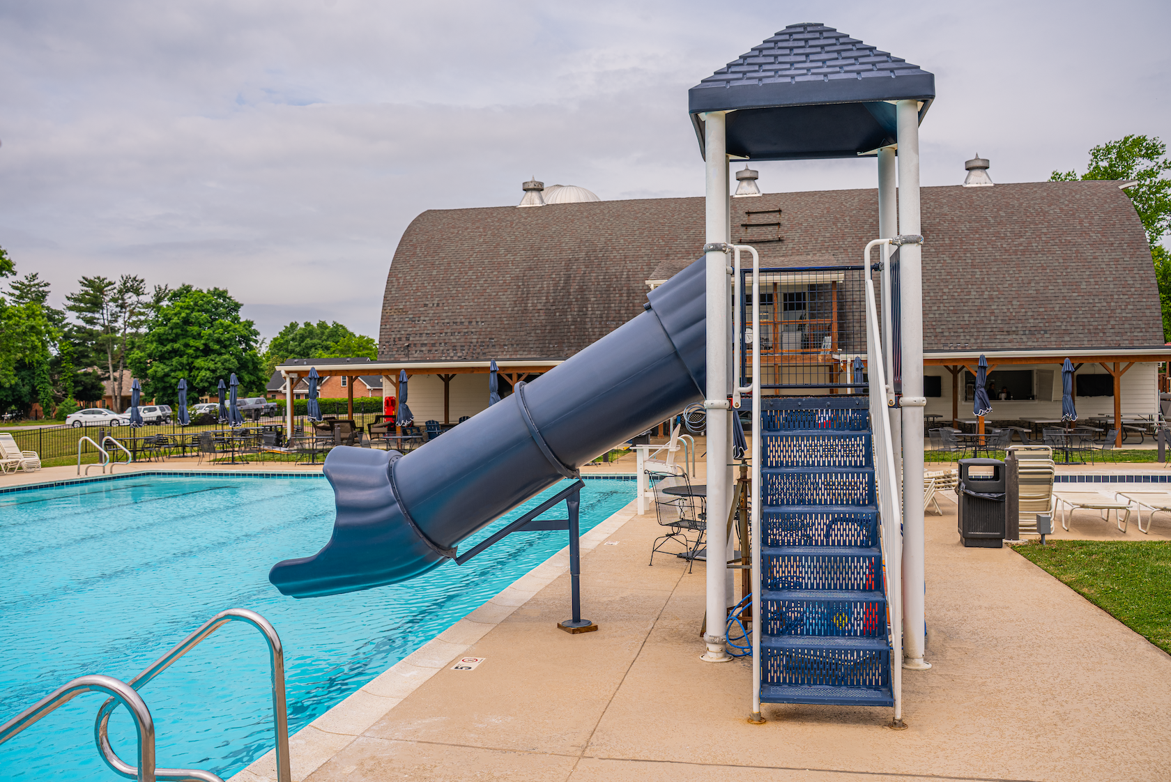 Community swimming pool with a blue slide, surrounded by lounge chairs, tables with umbrellas, and a building in the background with a brown roof and white walls.