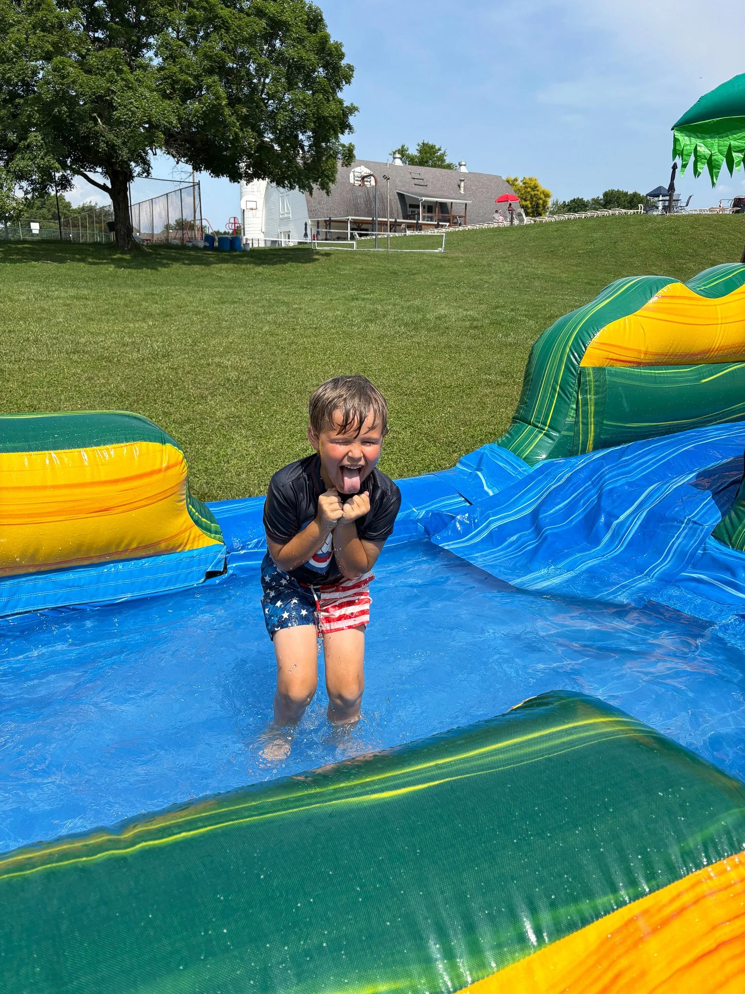 A young boy playing in an inflatable pool outdoors on a sunny day, wearing patriotic swim trunks, with a big smile and tongue out.