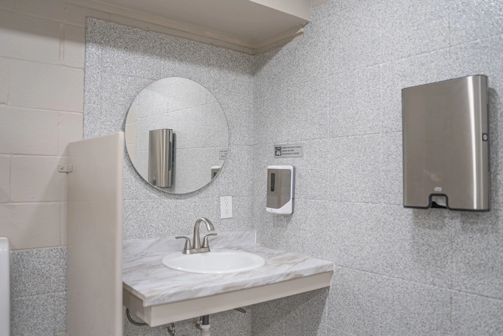 Public restroom sink area with a marble countertop, oval mirror, soap dispenser, and a paper towel dispenser on gray speckled wall.