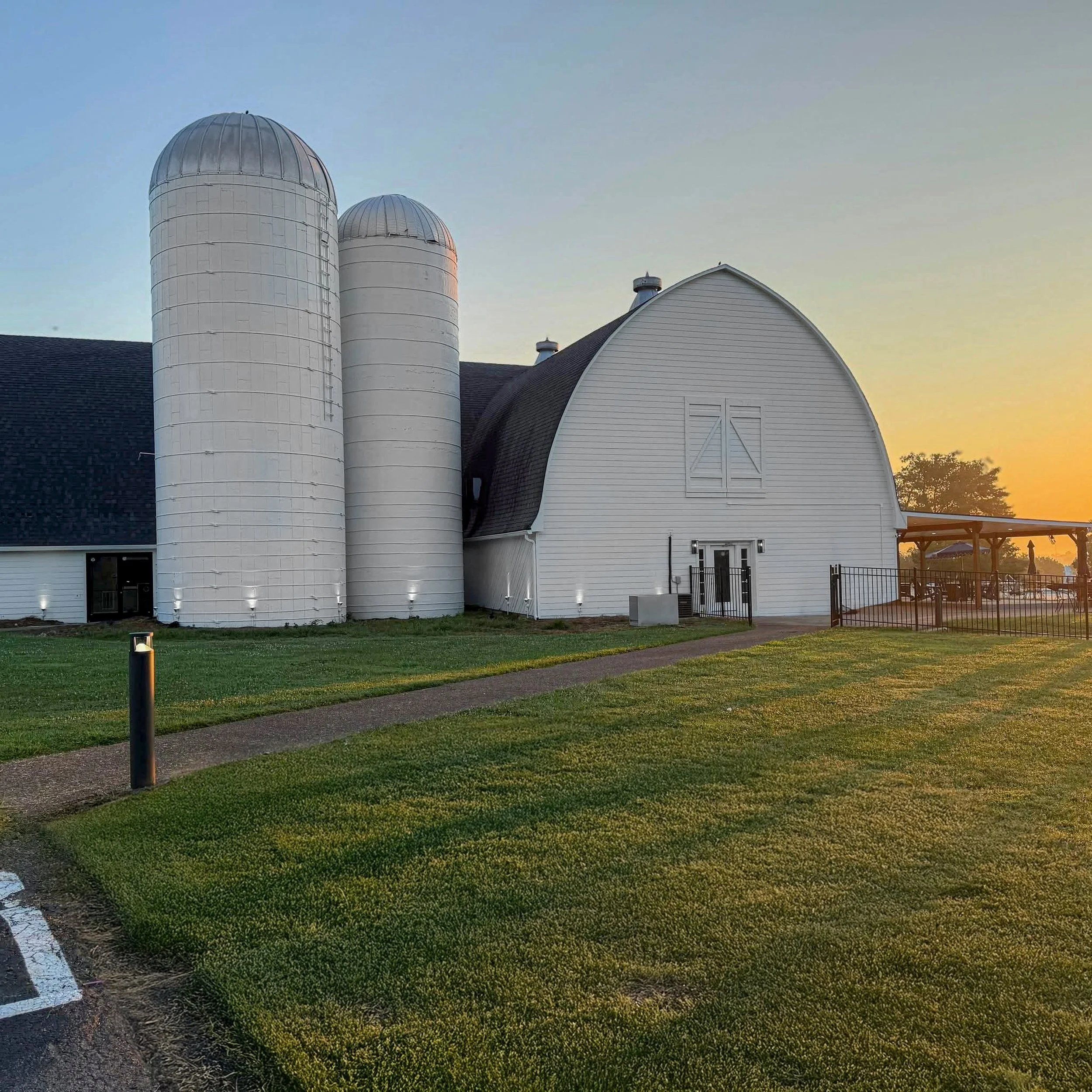 A white barn with two silos at sunset, green grass, a pathway, and a black fence.