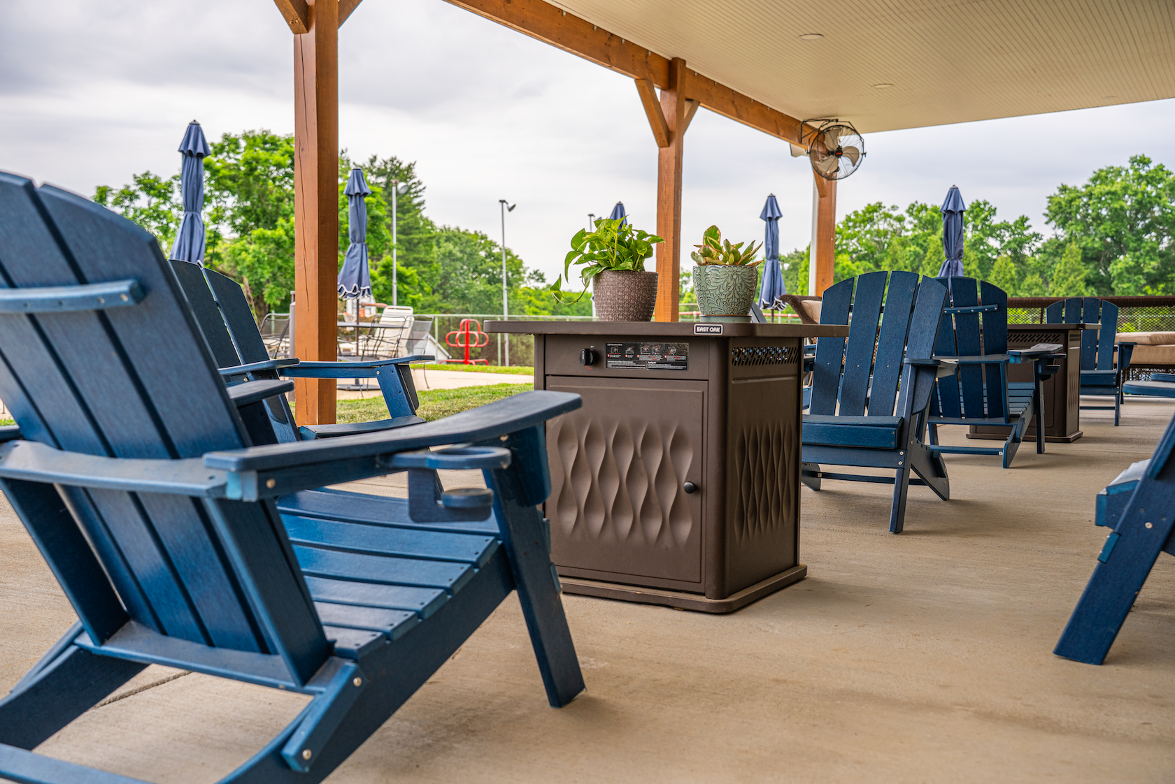 An outdoor patio area with blue Adirondack chairs, a brown heater, and potted plants, with a covered roof and trees in the background.